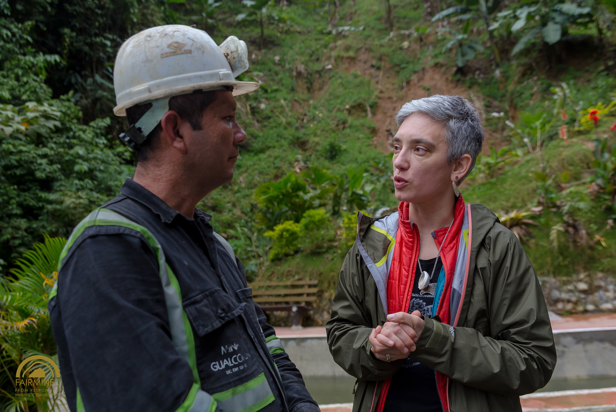 Two people talking outdoors, one with a white hard hat and safety vest, the other with short gray hair and a green jacket, surrounded by green plants and bushes.