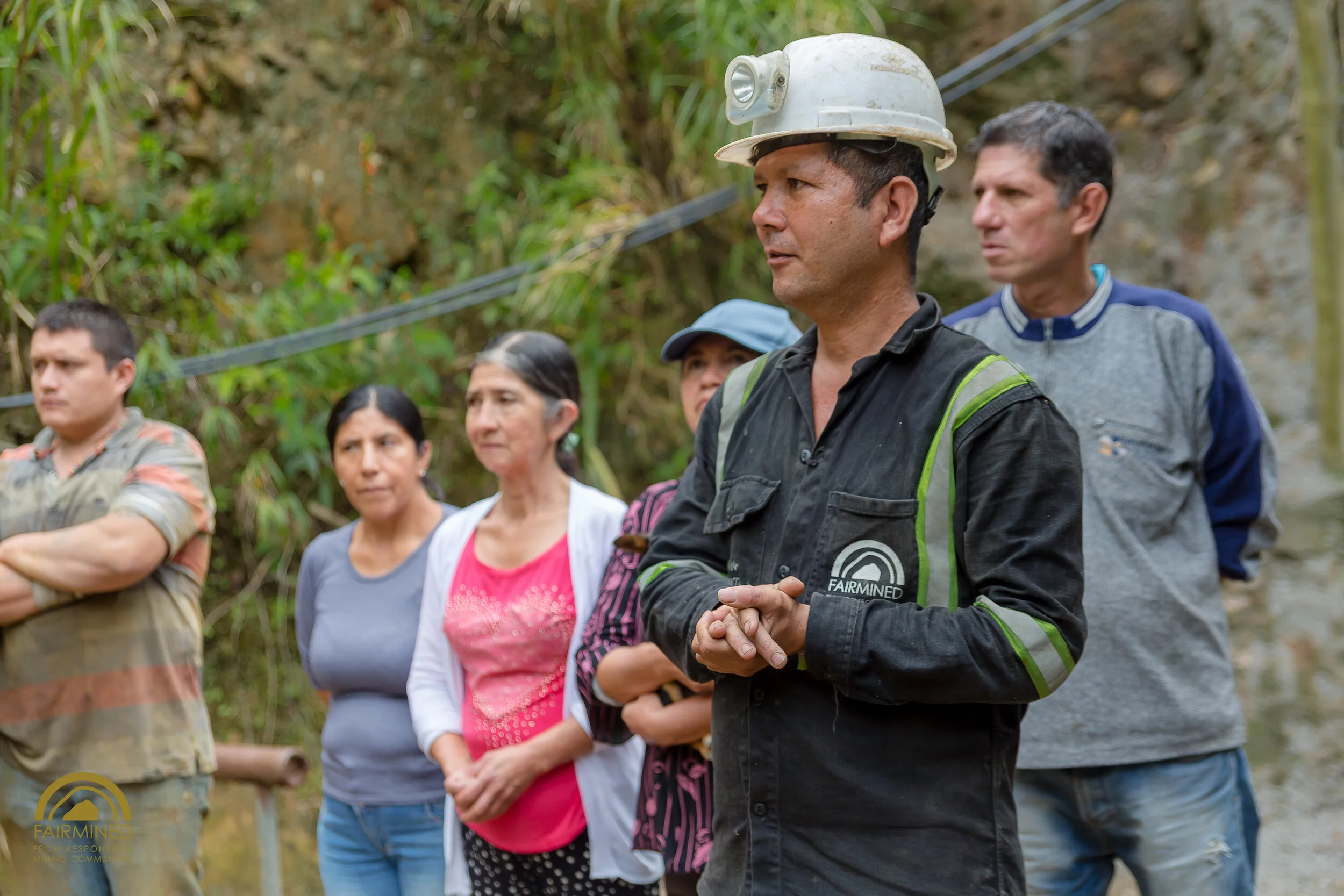 Group of six people standing outdoors against a wooded background, with one man in the foreground wearing a helmet and black shirt with 'Fairmined' logo, appearing to be speaking or listening