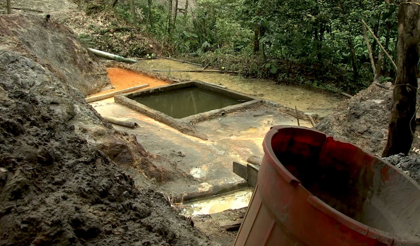 Construction site in a forest, with a rectangular concrete pool filled with dark water, surrounded by dirt and construction materials, including a large orange bucket in the foreground.