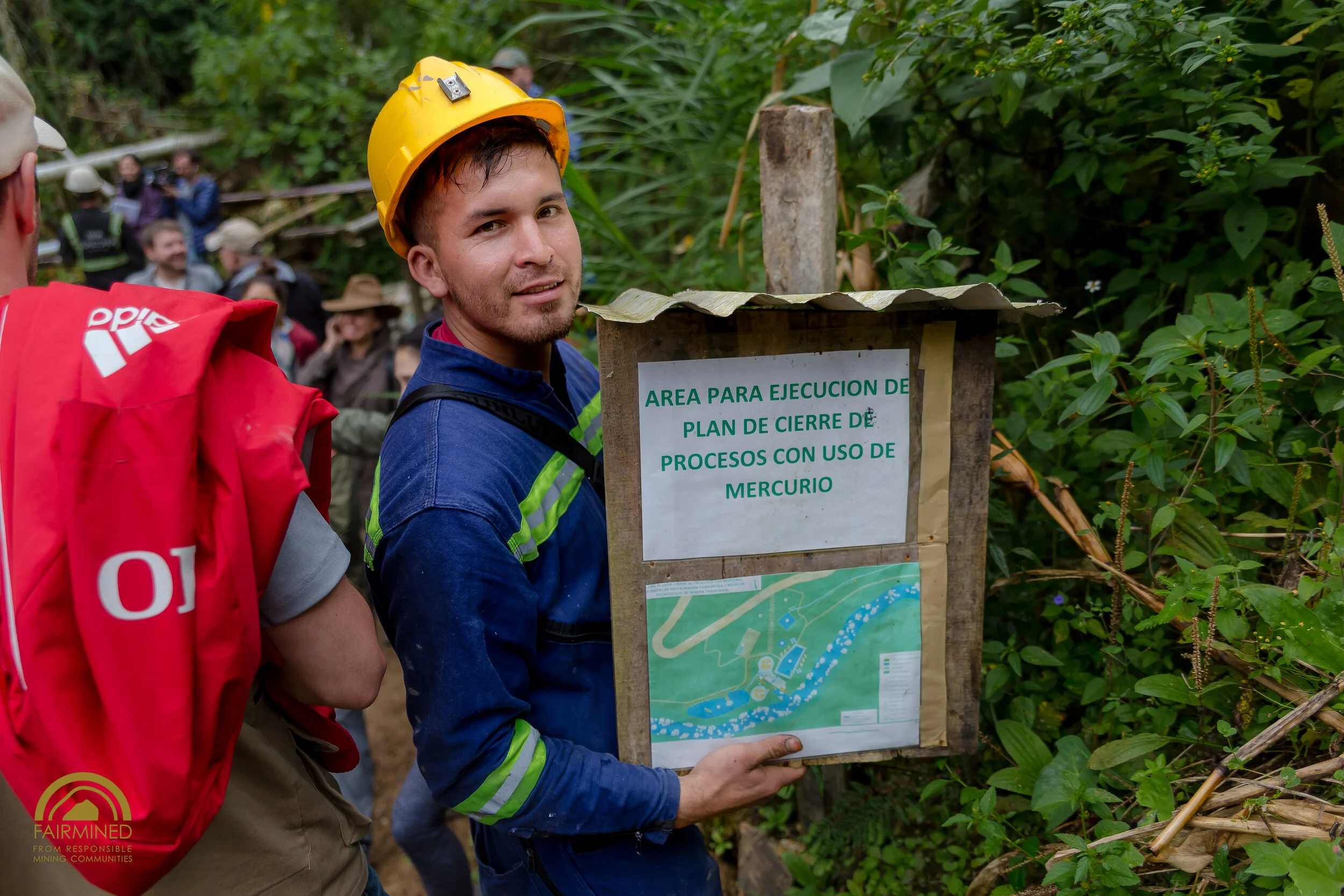 A man wearing a yellow safety helmet, a blue jacket with reflective stripes, and a backpack is standing in a lush green area holding a wooden sign with a map and Spanish text. Other people are visible in the background in a forested environment.
