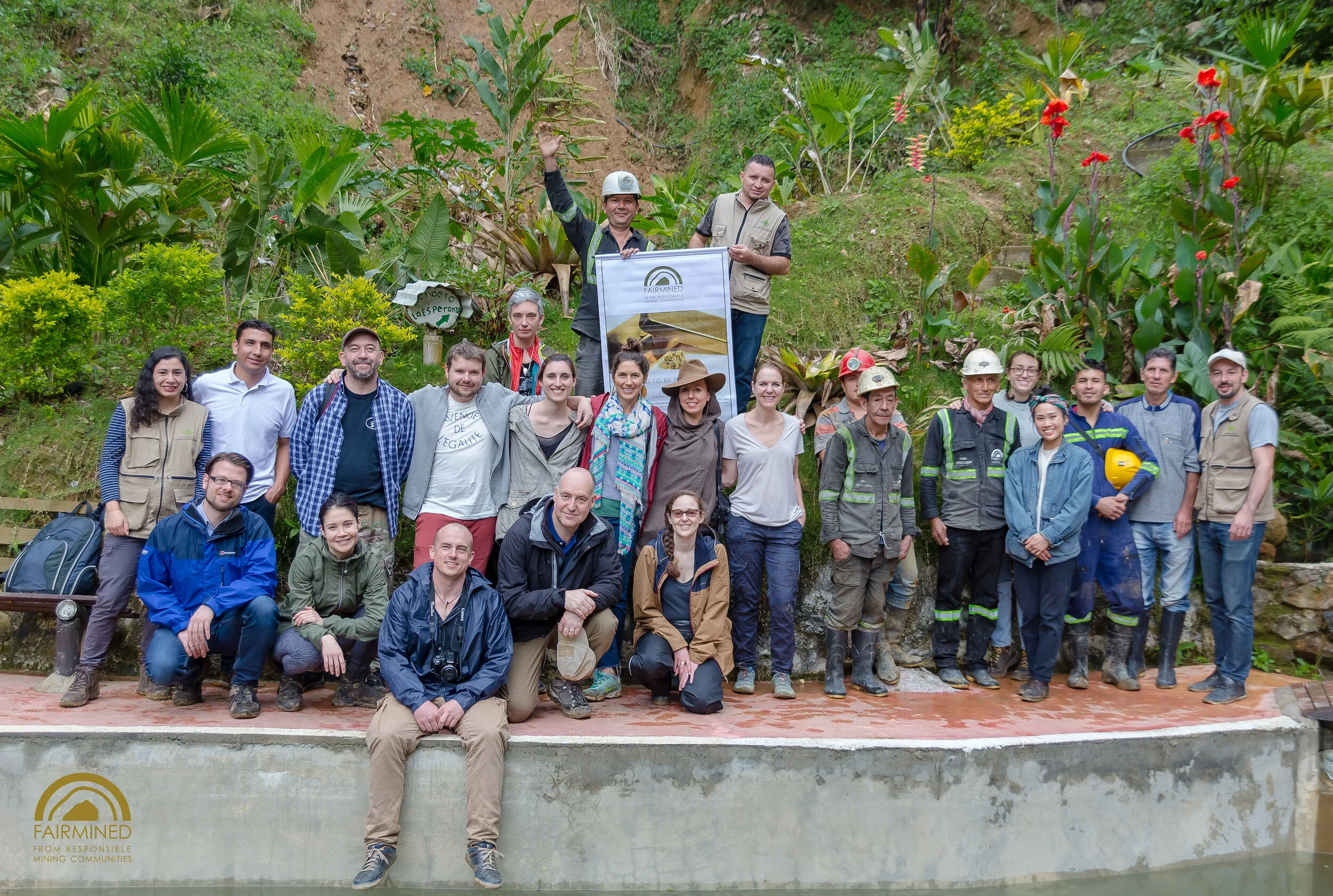 Group photo of people outdoors in front of lush green plants and trees, some wearing helmets and work uniforms, holding a banner with the FAIRMINED logo, during a community mining event.