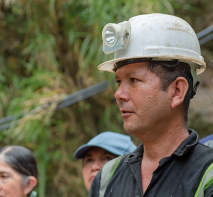 A man wearing a hard hat with a headlamp, outdoors with others in the background.