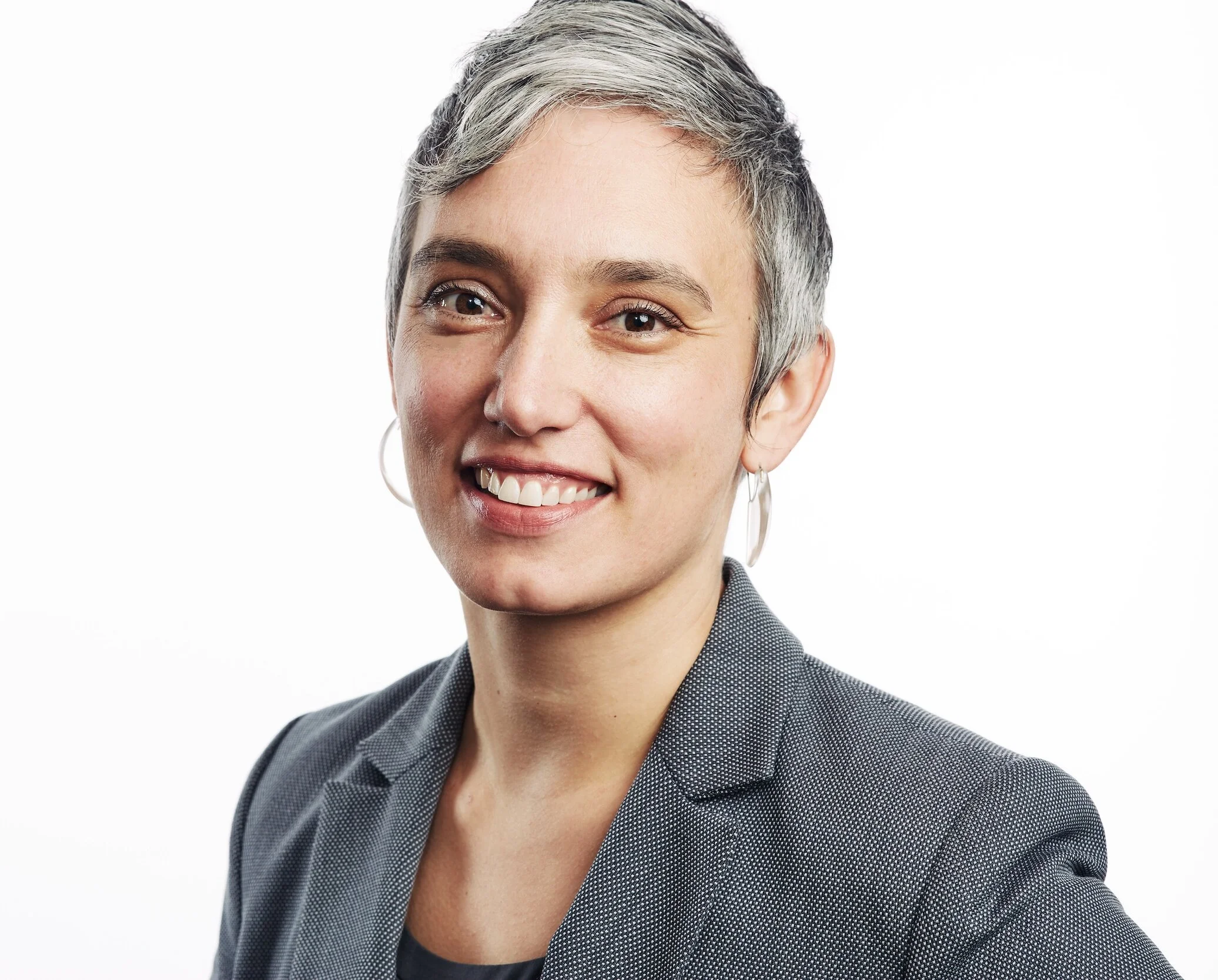 A woman with short, gray hair smiling, wearing a gray blazer and hoop earrings, against a plain white background.