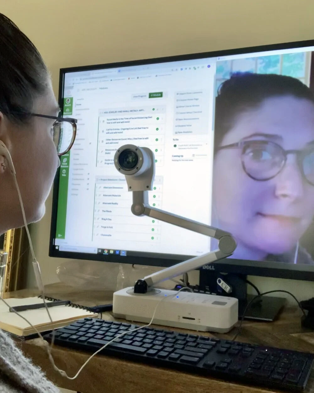 Person with glasses participating in a virtual meeting or telehealth session, looking at a computer monitor showing a woman’s face, with a camera mounted on a flexible arm directed at her face.