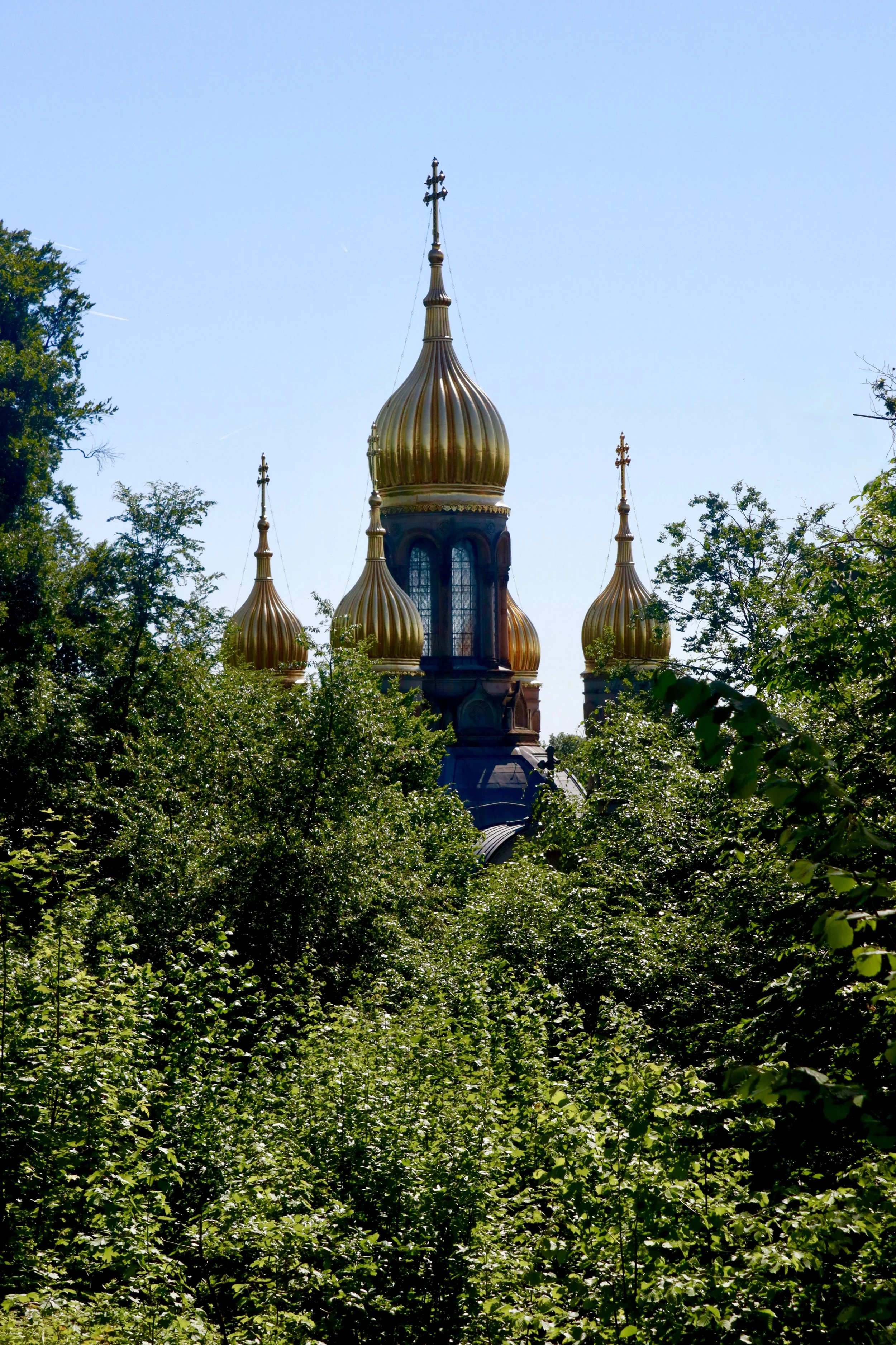Tall onion-shaped domes with crosses on top, part of a church, seen through lush green trees against a bright blue sky.