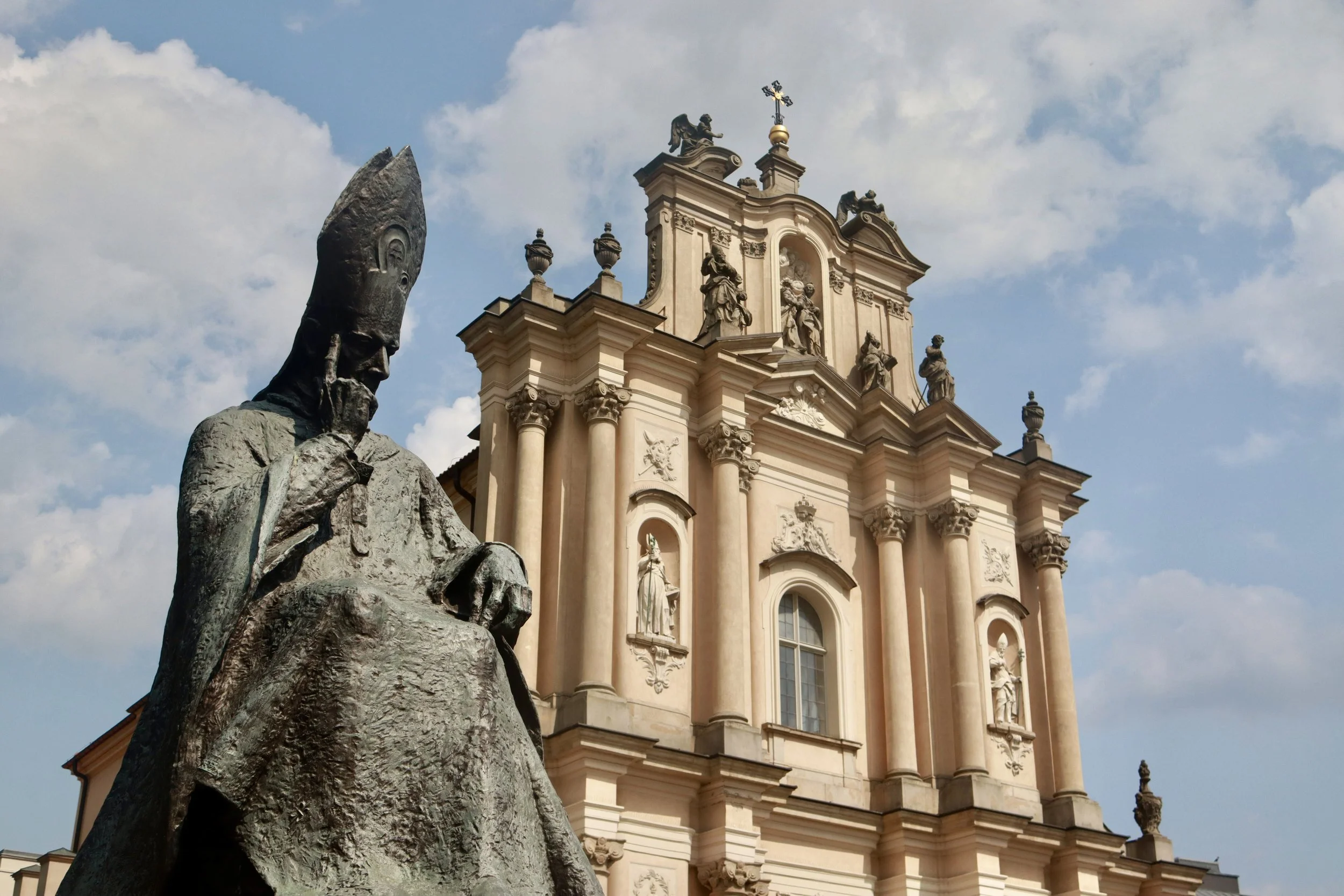 Statue of a religious figure in front of a baroque church with ornate sculptures and decorative elements, under a partly cloudy sky.