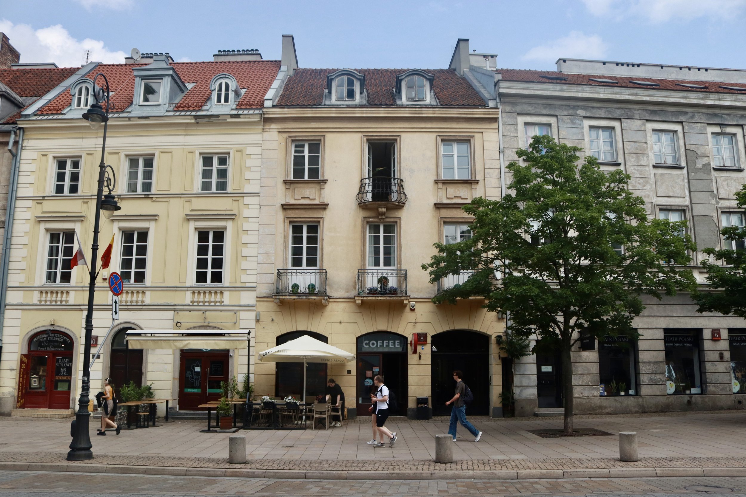 European street scene with colorful historic buildings, outdoor café with tables and umbrellas, pedestrians walking, trees, and street lamp.