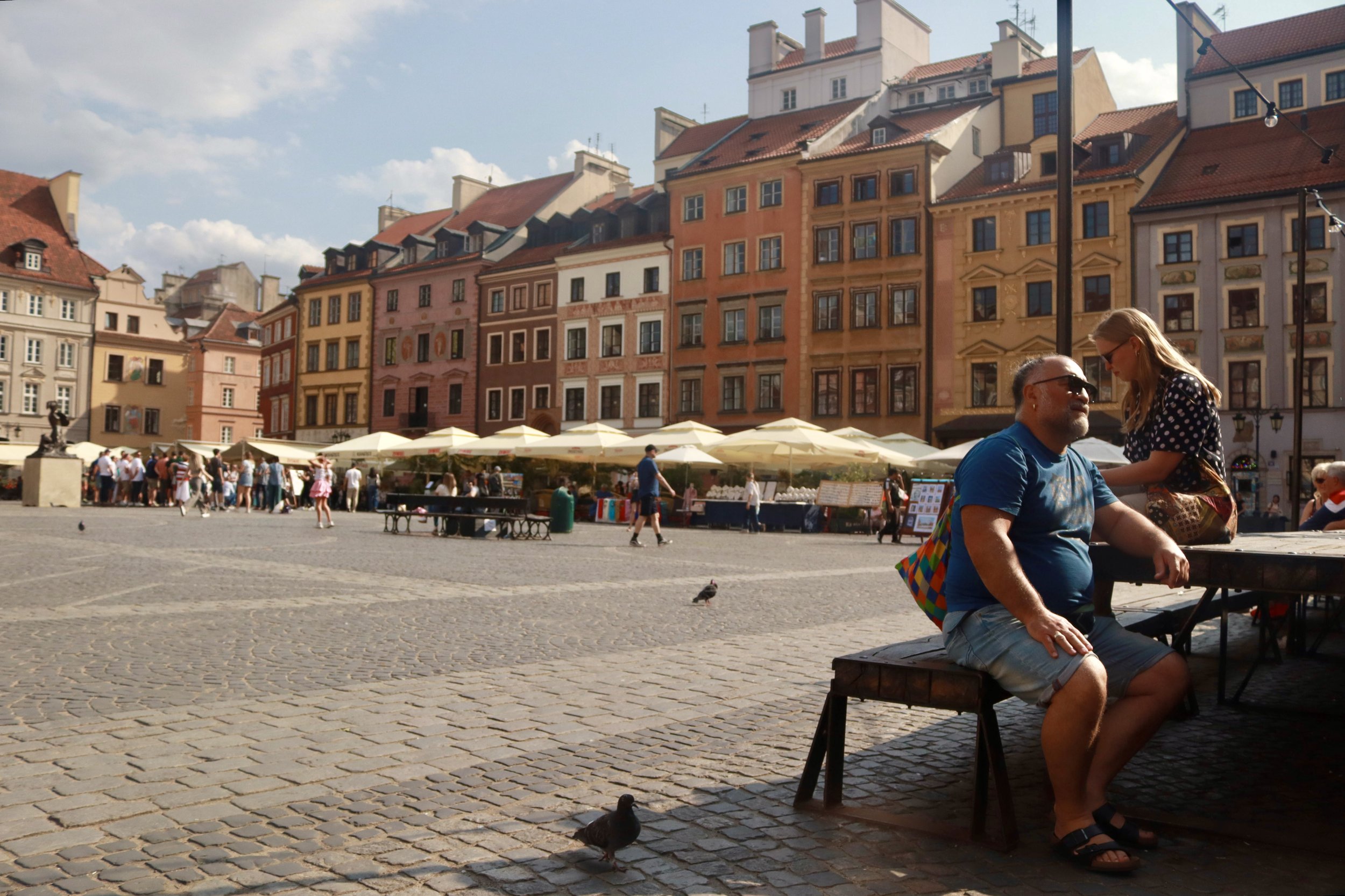 A city square with colorful buildings, outdoor market stalls under umbrellas, and people walking or sitting at outdoor tables. Two people are seated at a table in the foreground, with a man in a blue shirt and a woman leaning over him. A pigeon is on