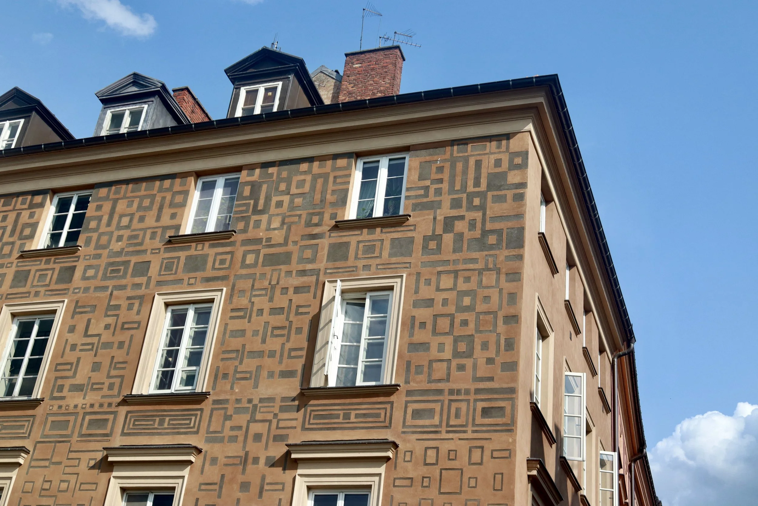Close-up of a building with geometric patterned facade, multiple white-framed windows, and a dark roof with dormer windows against a blue sky.