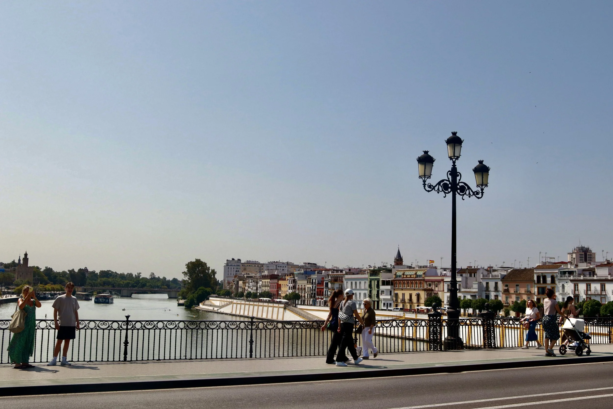 People walking along a riverside promenade with buildings and a bridge in the background, a decorative street lamp, and a clear sky