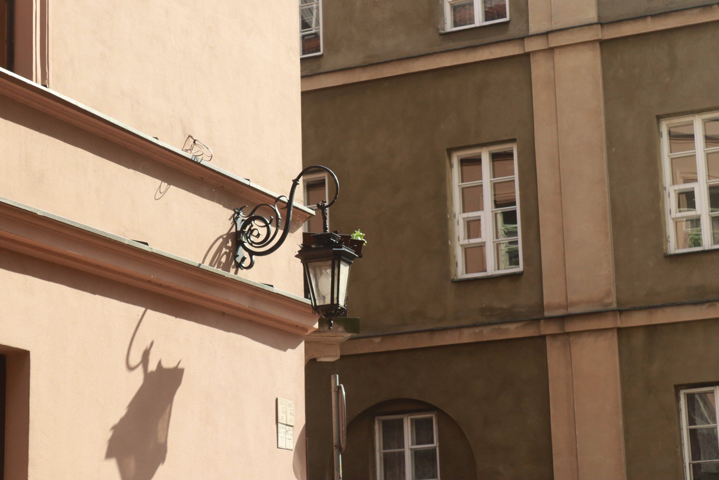 A street corner with pink and beige buildings, a vintage-style street lamp, and shadows cast on the wall.