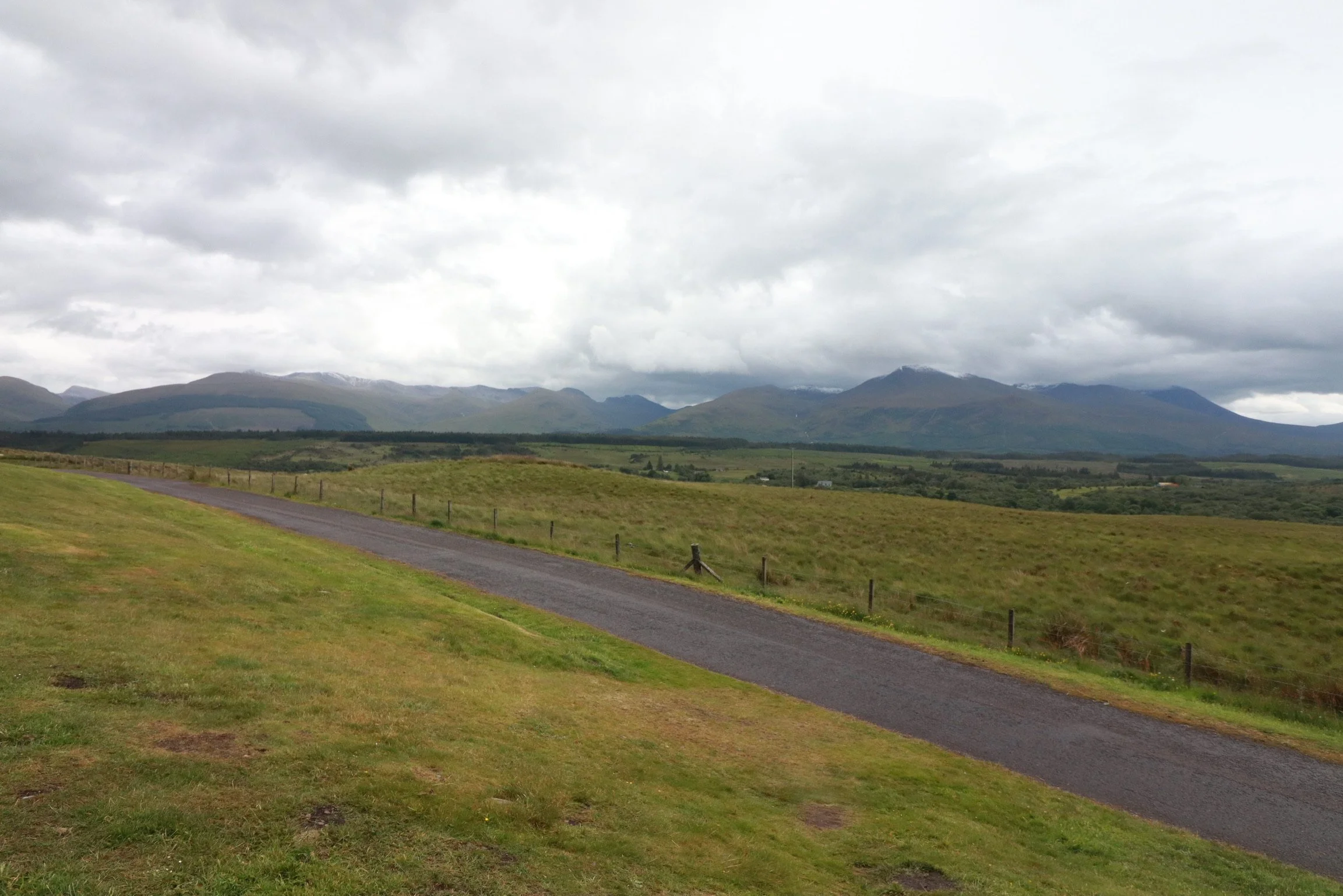 Scenic landscape with a grassy field, a narrow asphalt road, rolling hills, and mountains in the distance under a cloudy sky.