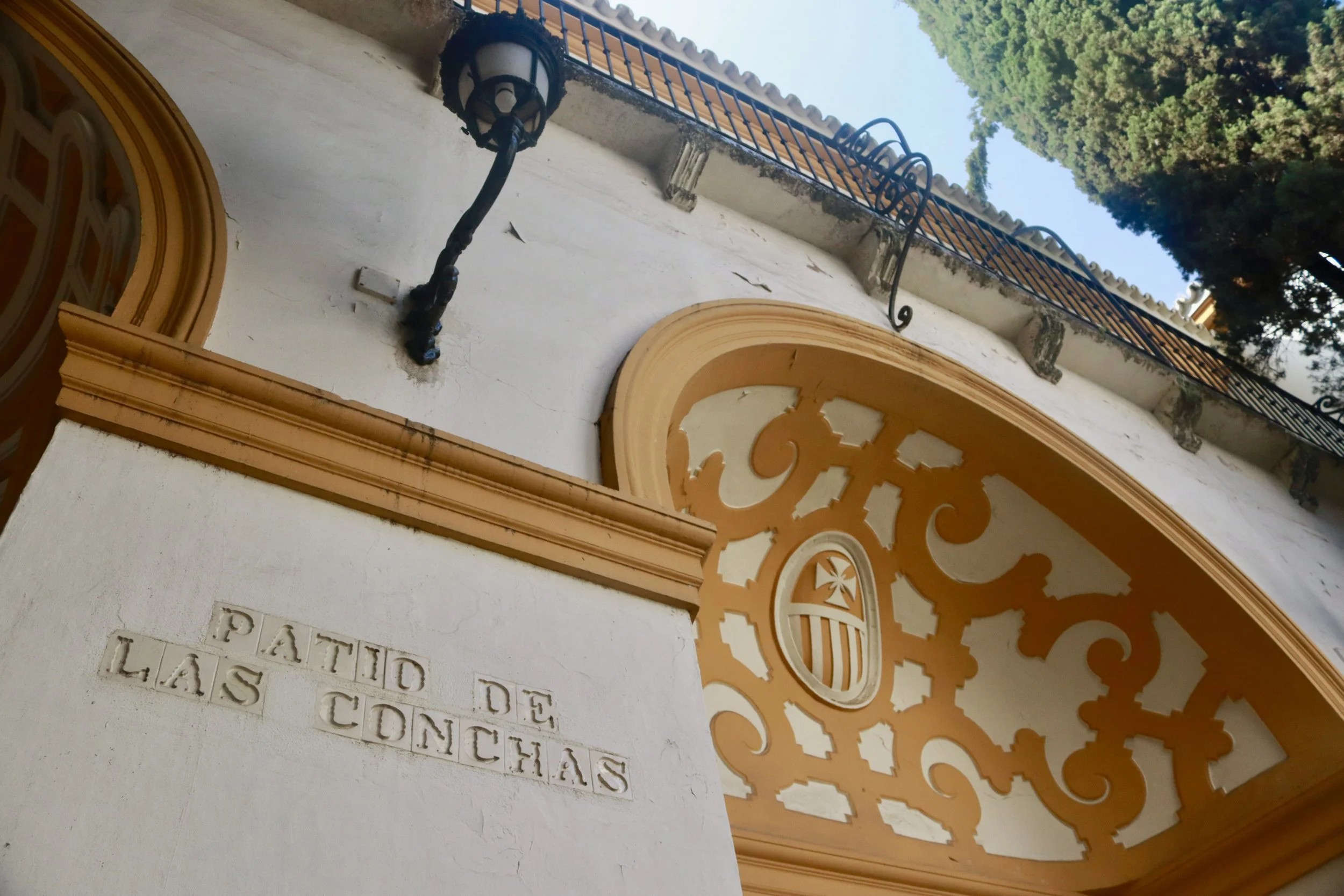 Historical building corner with ornate decorative orange archway, a black wrought iron street lamp, and a sign reading 'Patio de Las Conchas' in Spanish, with trees in the background.