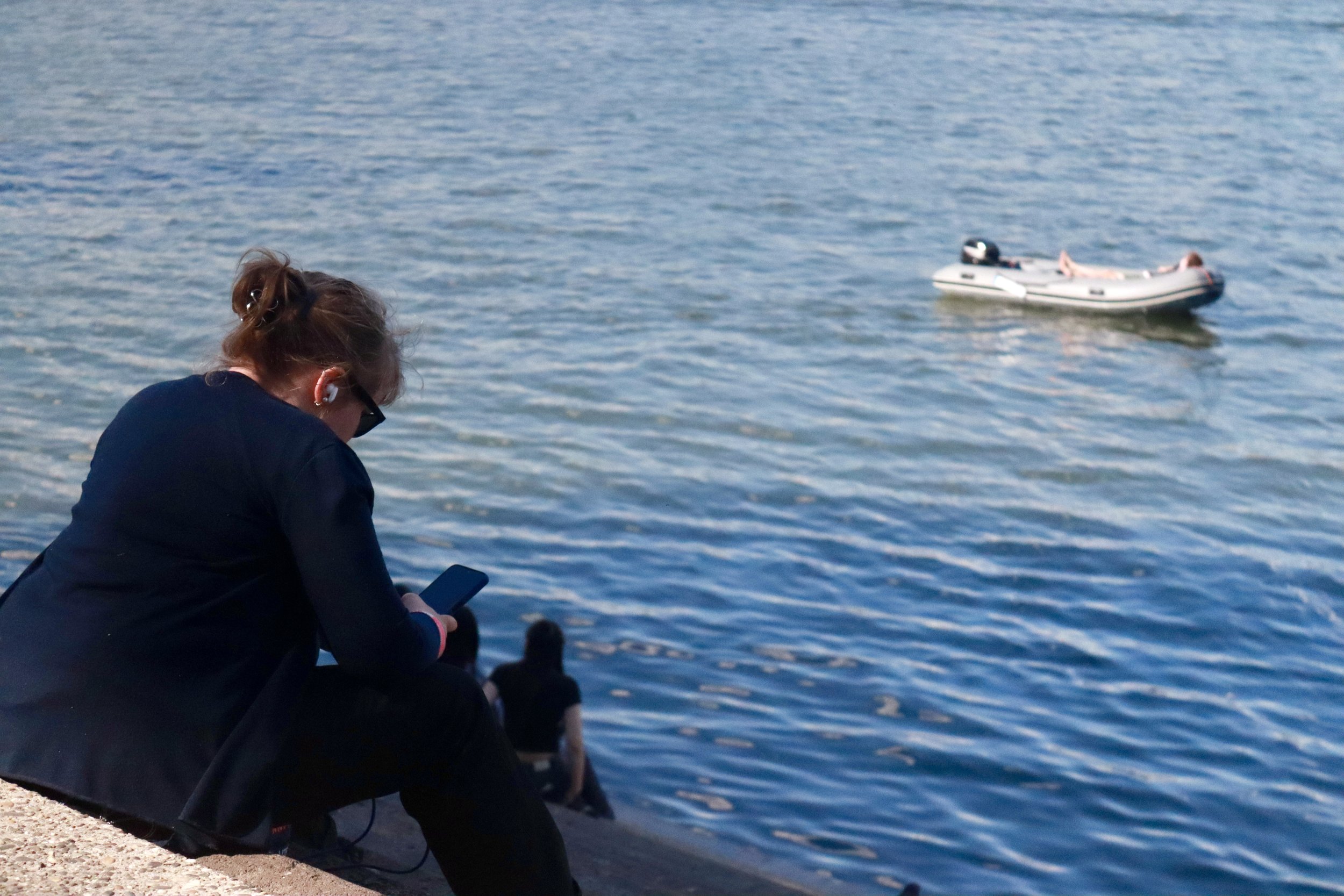 A woman with earbuds and sunglasses sitting on a concrete edge by the water, looking at her phone, with a small boat floating in the water nearby.