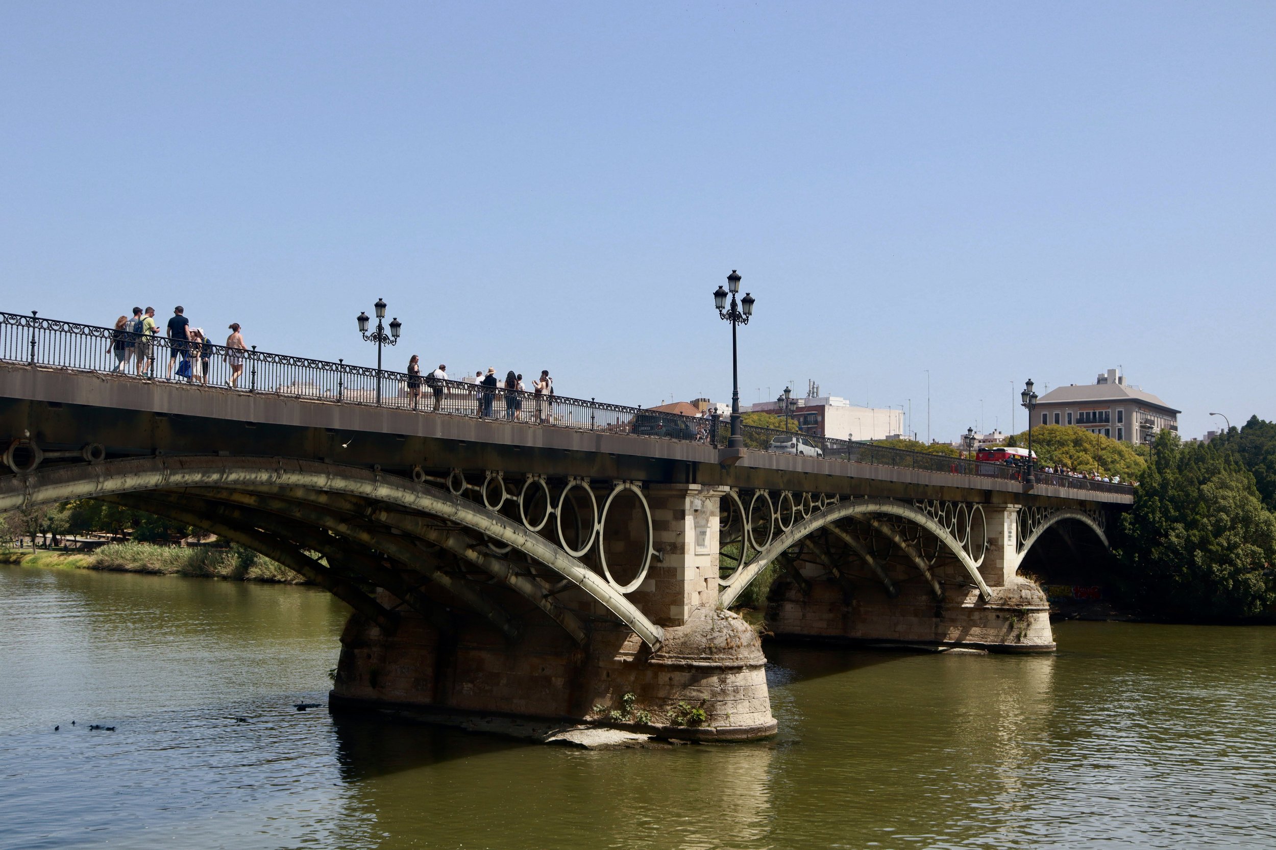 People walking across a bridge over a body of water on a clear day, with buildings and trees in the background.