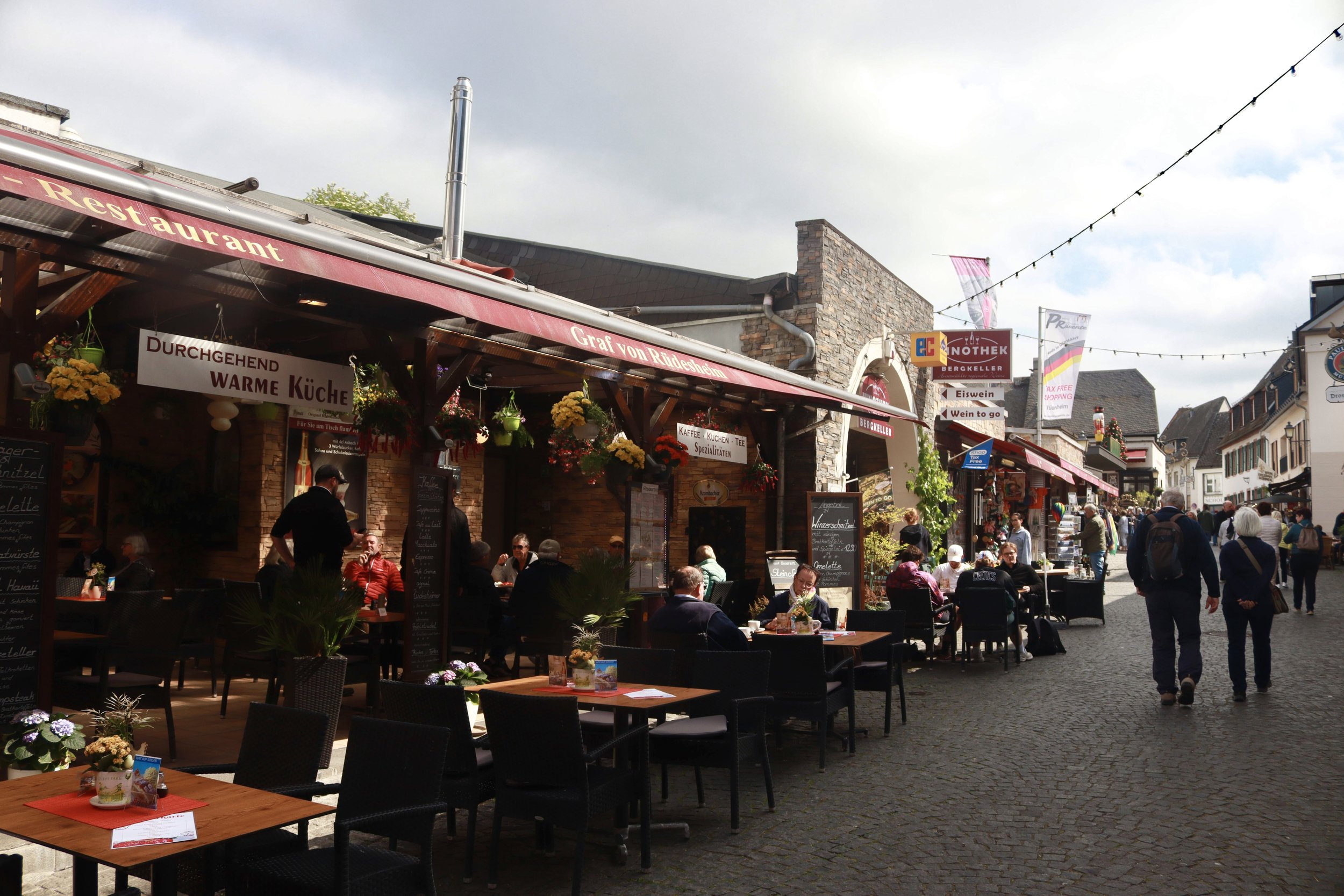 Outdoor cafe with tables and chairs on a cobblestone street, people dining and walking, storefronts with signs, and string lights overhead in a European town.