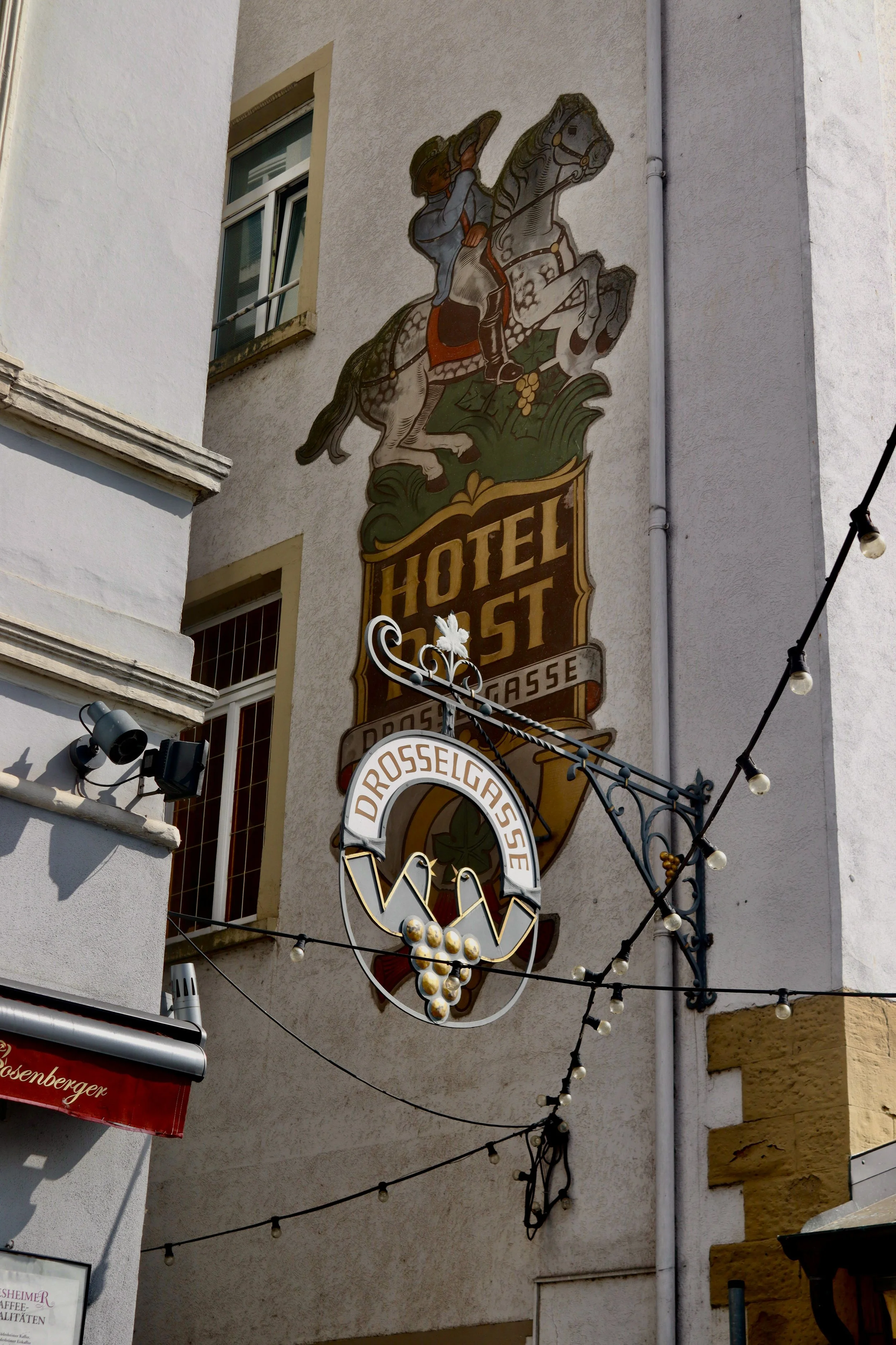 A mural on a building wall features a knight riding a horse, with a sign below that reads 'Hotel Gaff' and another sign with a bunch of grapes, indicating the location as 'Drosselgasse'.