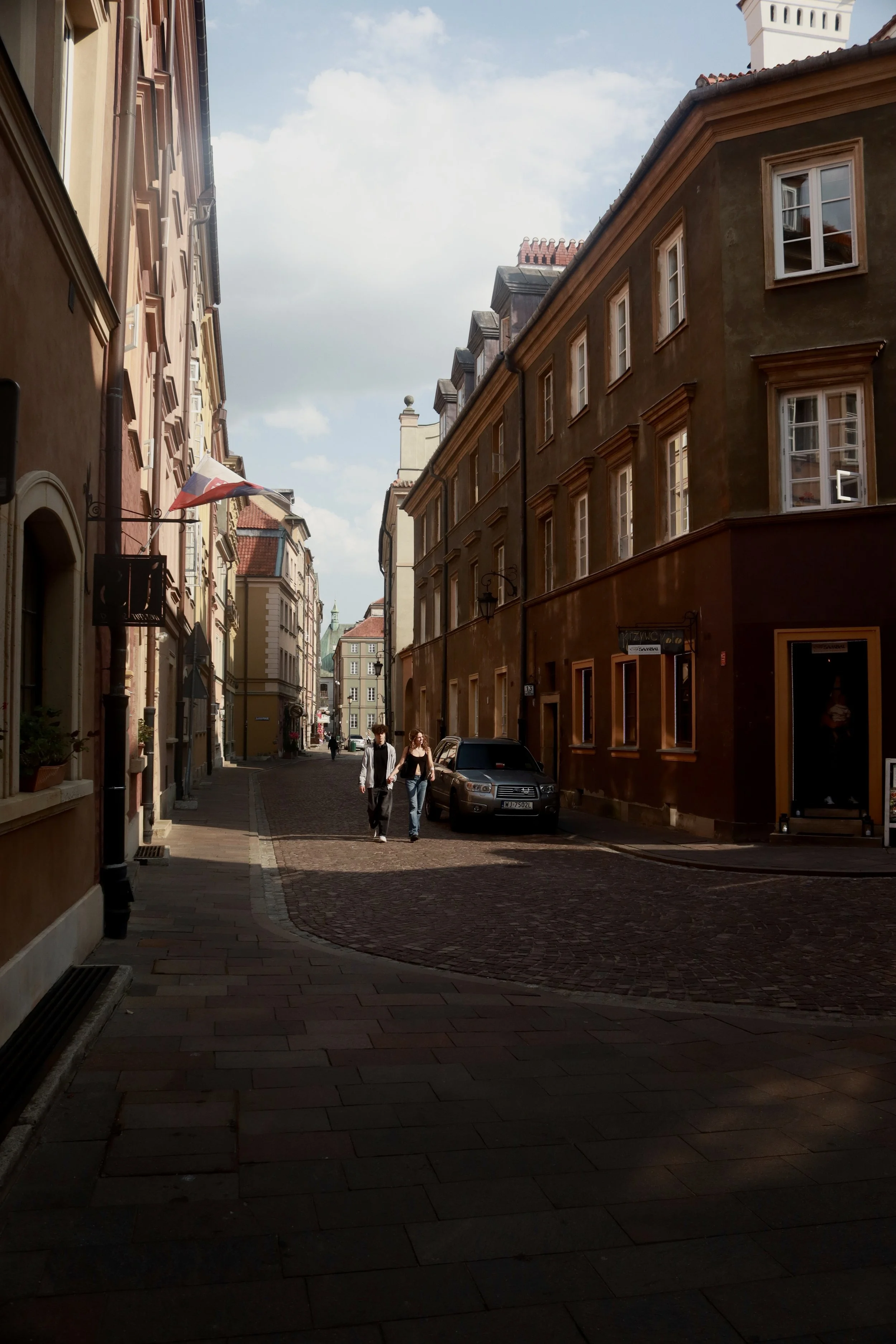 A narrow European street with cobblestone pavement, lined with historic buildings featuring colorful facades, and a few people walking in the daytime under partly cloudy skies.