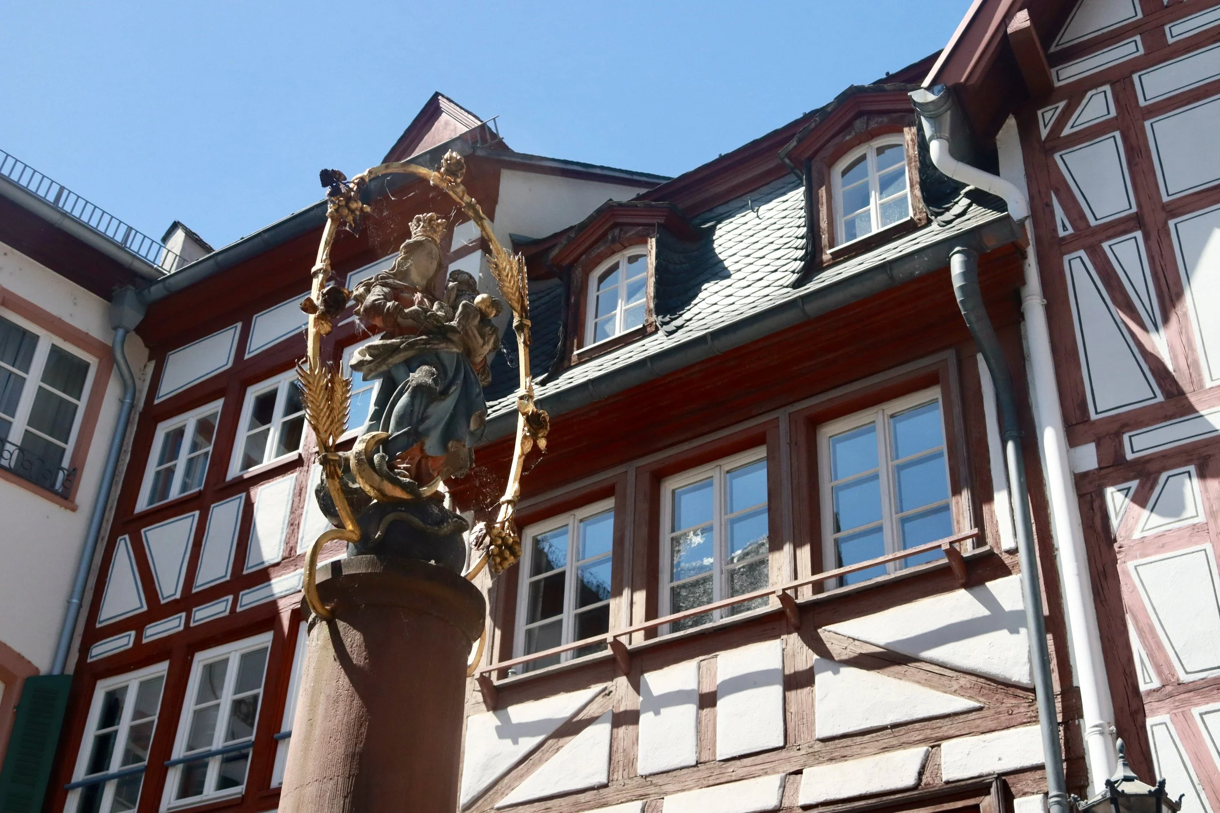 Building with traditional European architecture, featuring a decorative fountain with a religious sculpture on a column in the foreground.