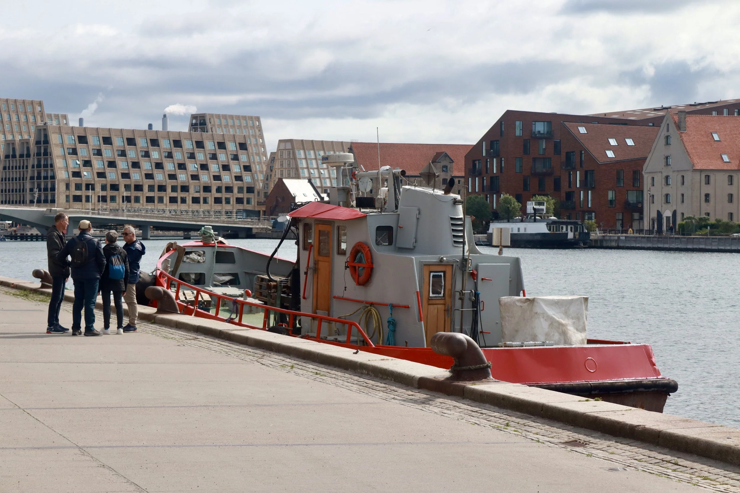 Four people standing near a red and gray tugboat docked at a harbor with modern and historic buildings in the background.