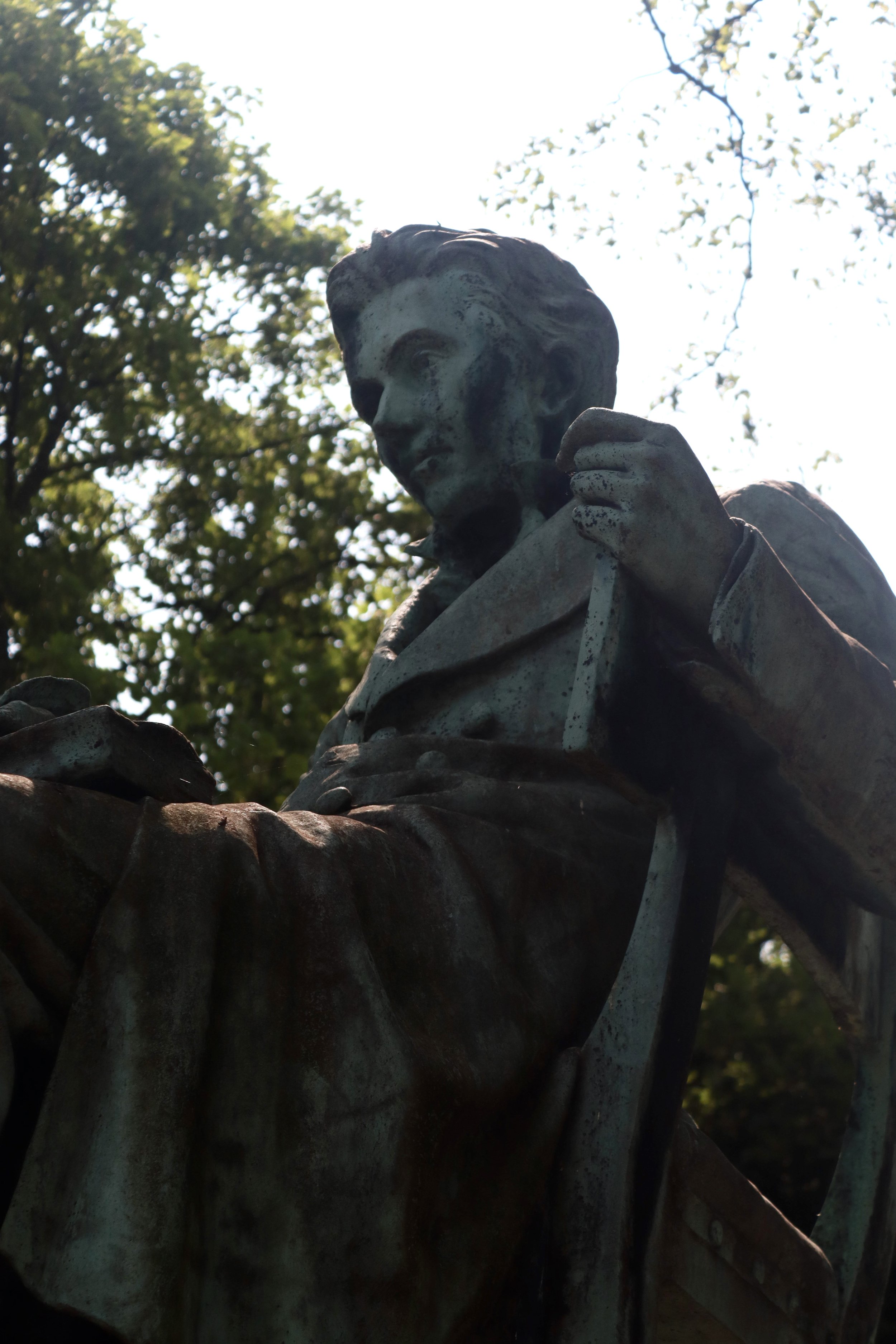 Close-up of a weathered bronze statue of a woman with short hair, wearing a coat, holding a flag or banner, against a backdrop of trees and a bright sky.