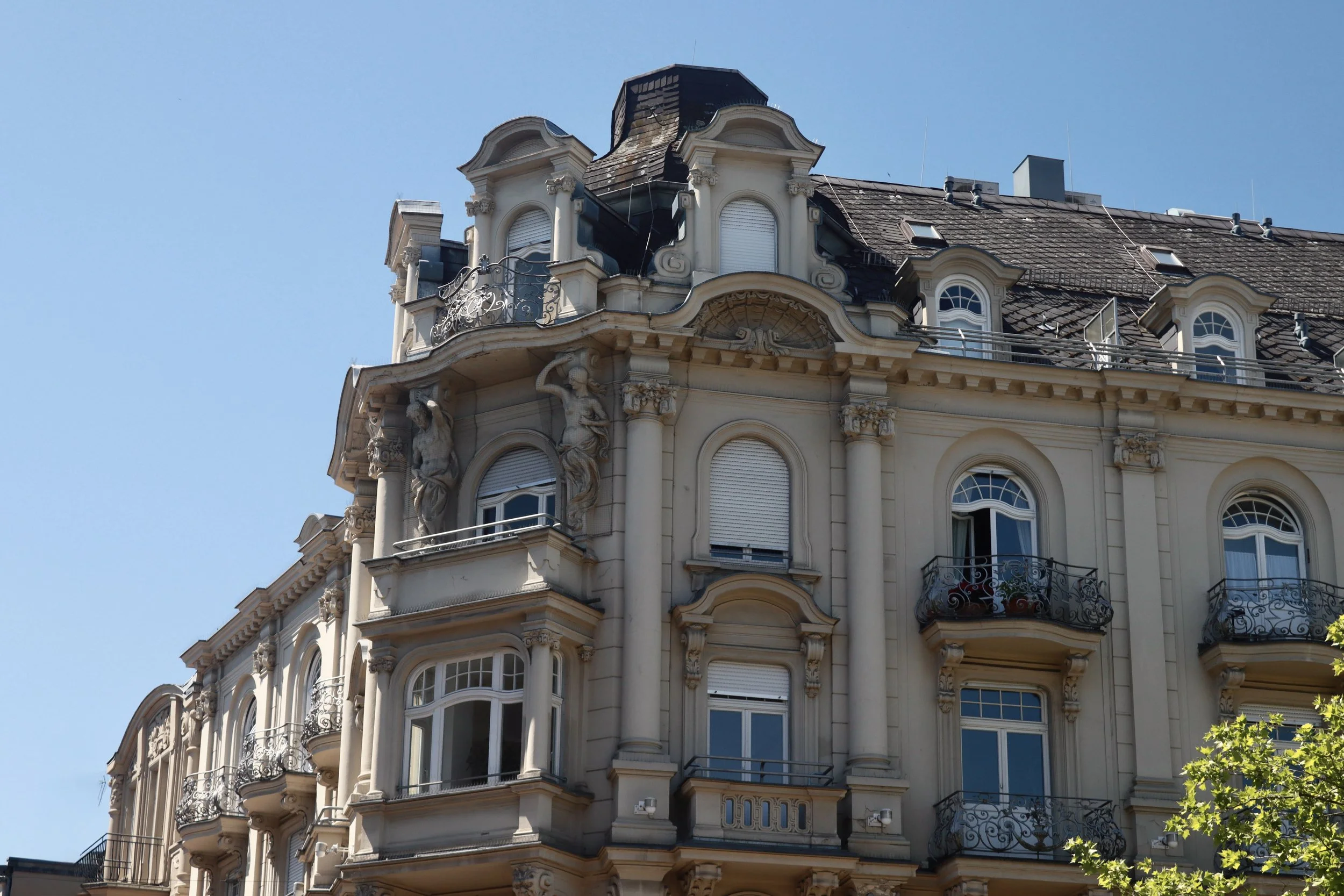 A historic multi-story building with ornate architectural details, balconies with decorative wrought iron railings, and sculpted figures on the facade, under a blue sky.
