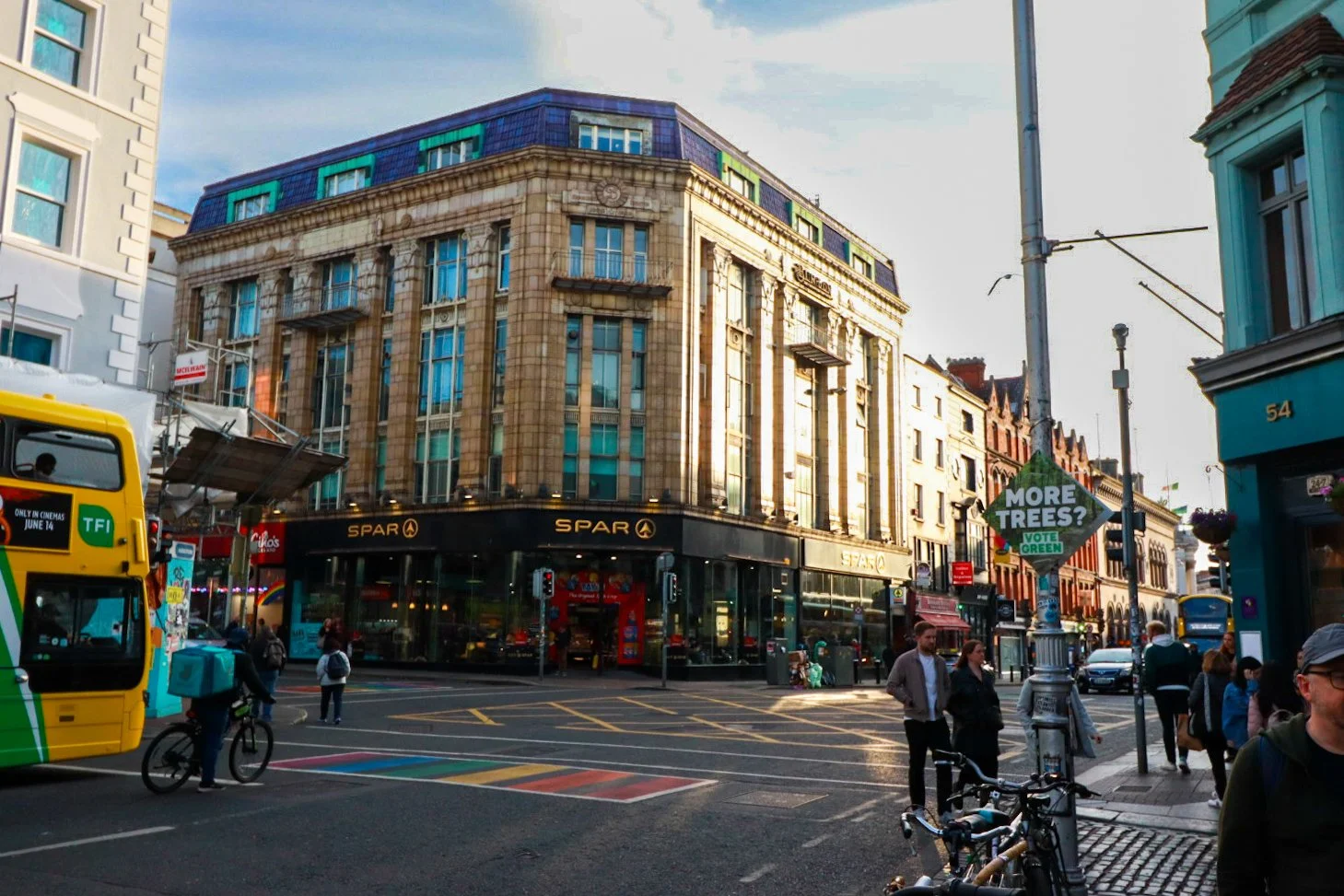 A busy city street corner with pedestrians, bicycles, and a double-decker bus. Buildings include a SPAR grocery store, with a sign asking "More Trees? Vote Green." The weather appears clear and sunny.