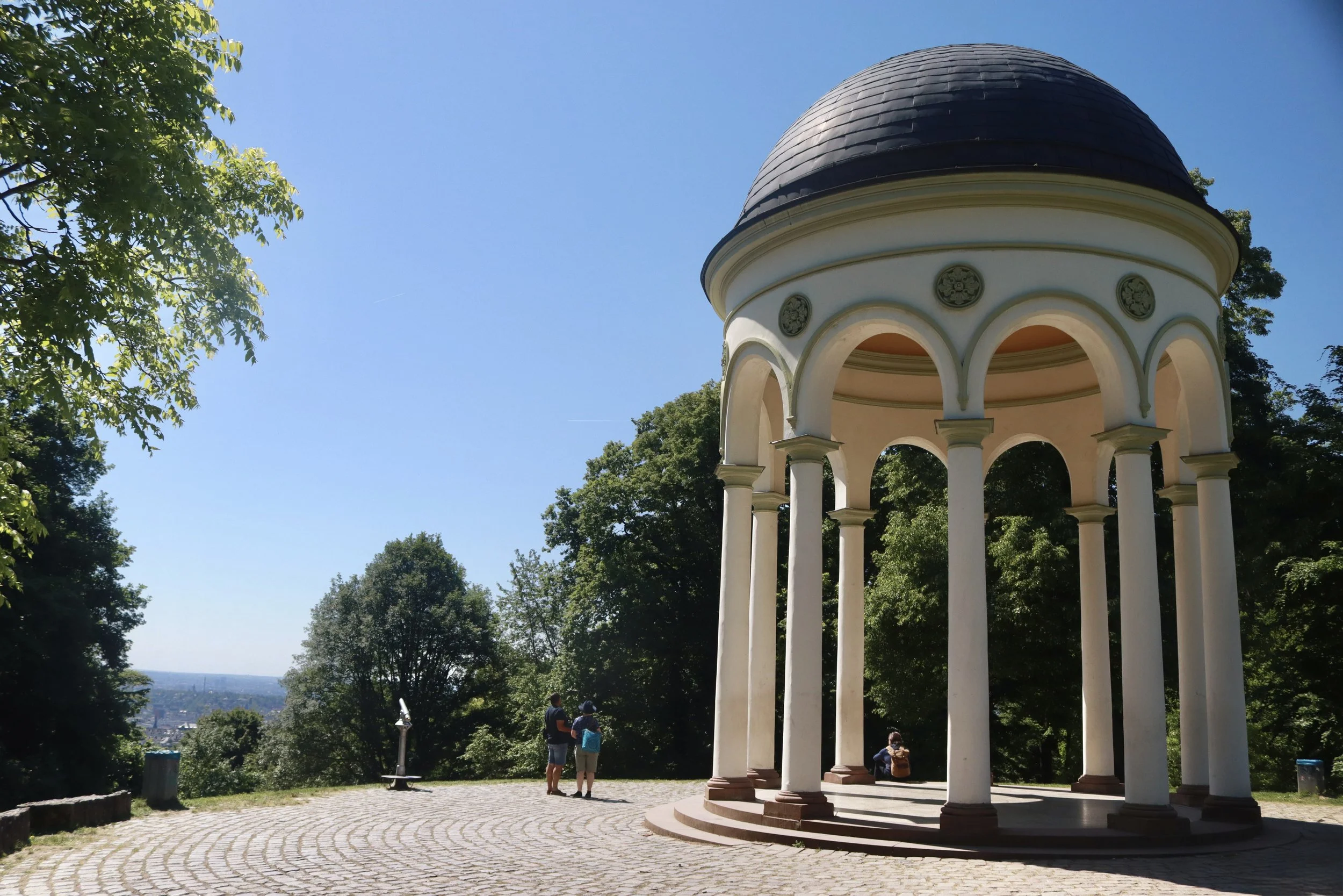 A small, white, circular pavilion with columns and a dome on a hilltop, overlooking a city and surrounded by green trees on a sunny day.