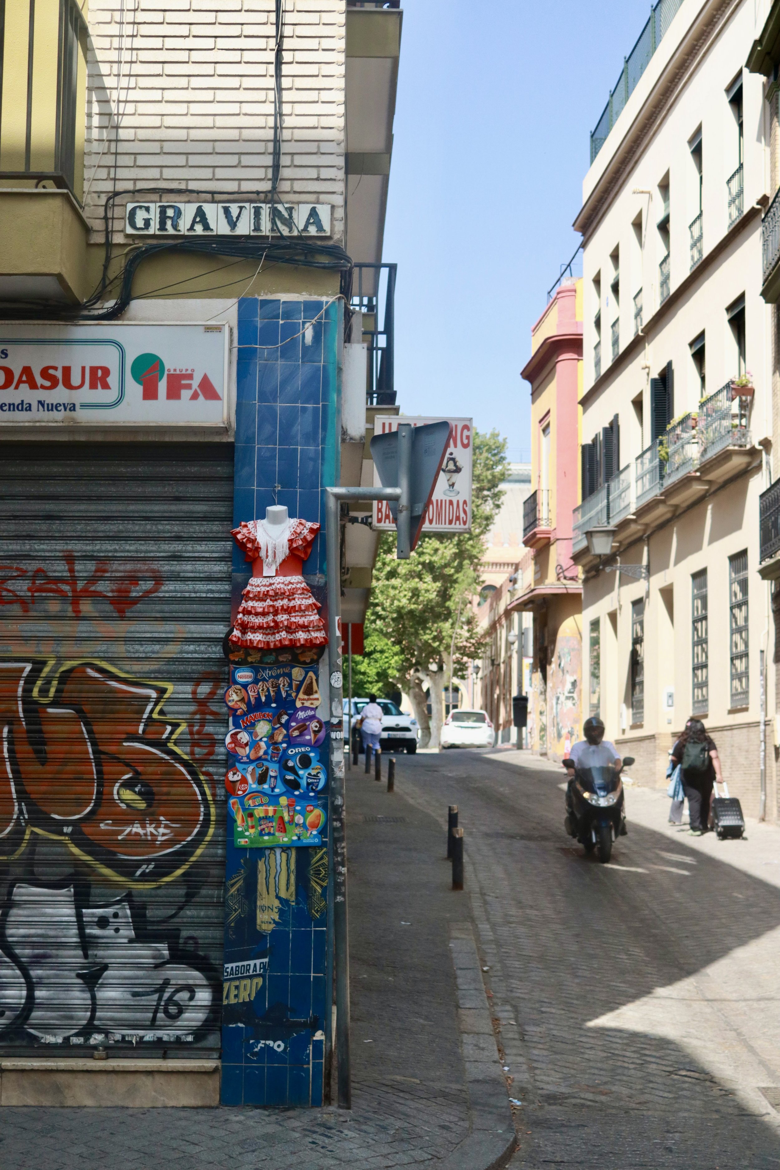 A street scene in a city with a narrow road, pedestrians, and a scooter. Buildings with balconies are visible, and a sign reads 'GRAVINA'. There's a decorated dress on a mannequin next to a wall covered in graffiti and ice cream posters.