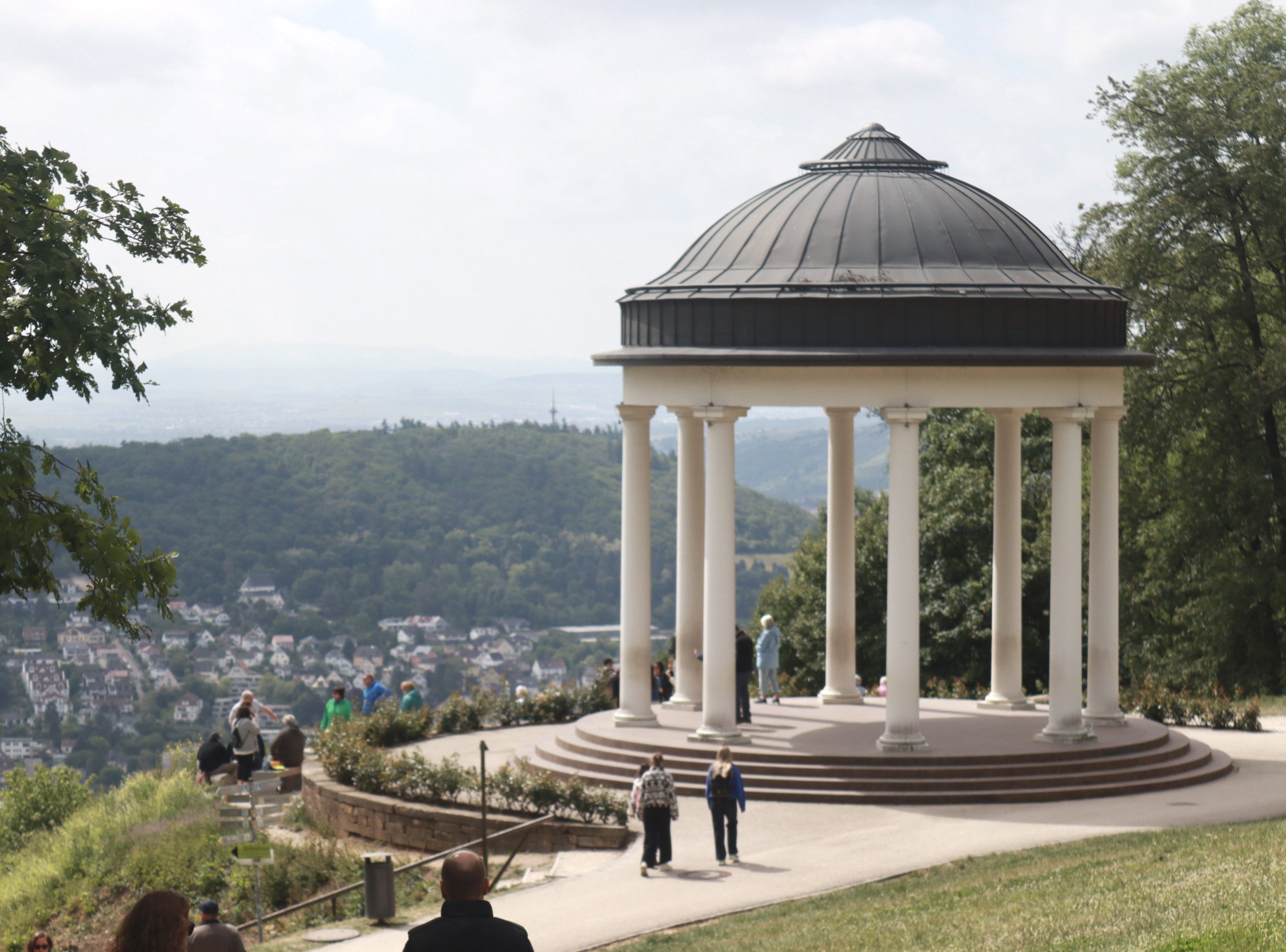 A white circular pavilion with columns and a domed roof on a hill, overlooking a town and green hills in the distance, with people walking and sitting around the area.