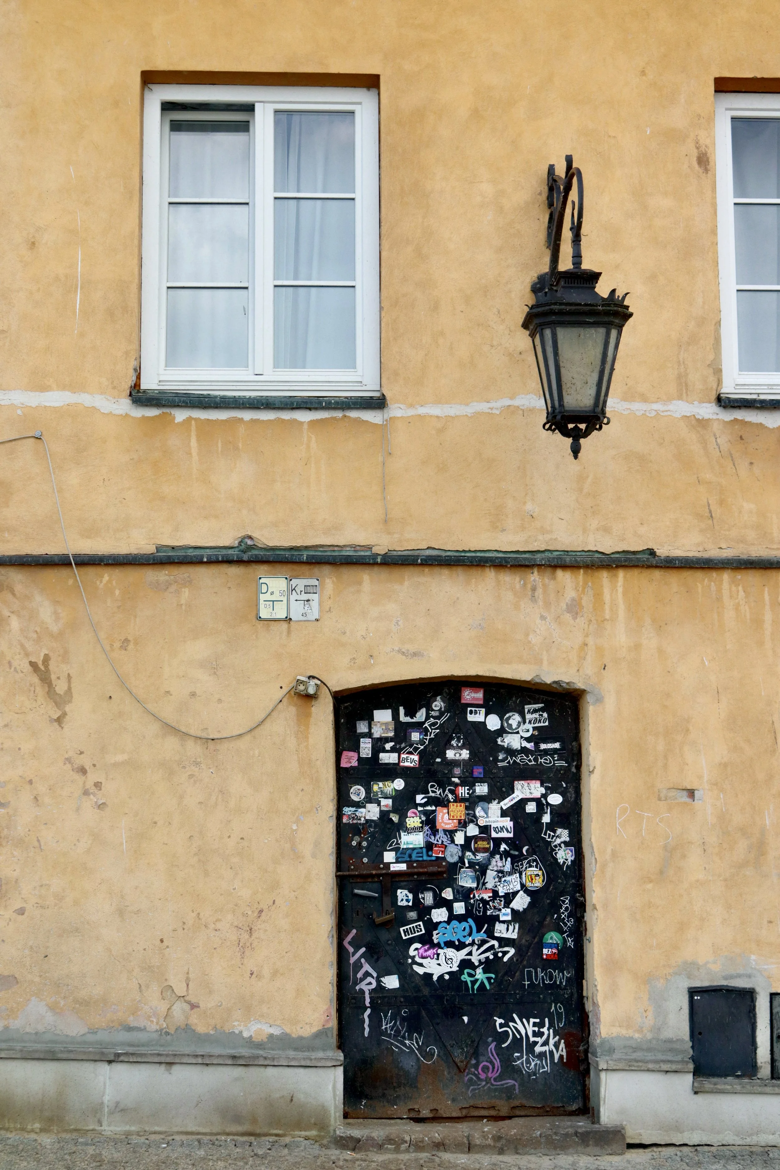 An old building facade with two white-framed windows, a vintage mounted street lamp, and a black door covered with numerous stickers and graffiti.