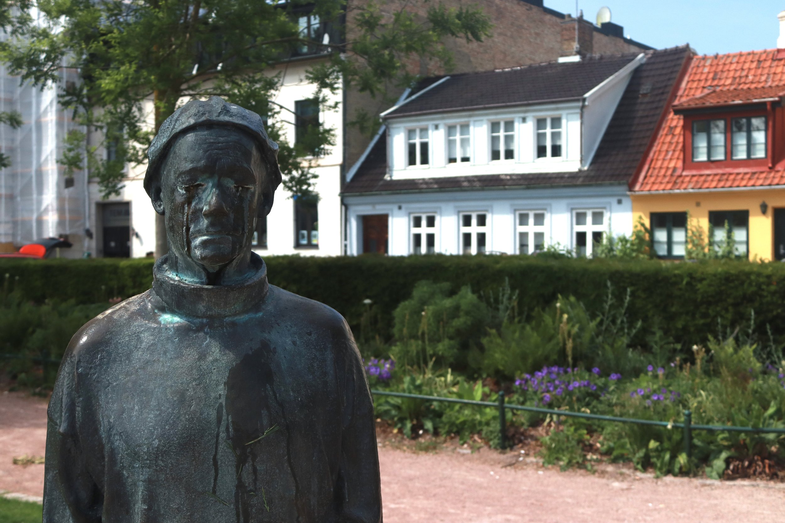 Bronze statue of a woman with a sad expression, standing outdoors in a park, with a background of colorful houses and a garden area.