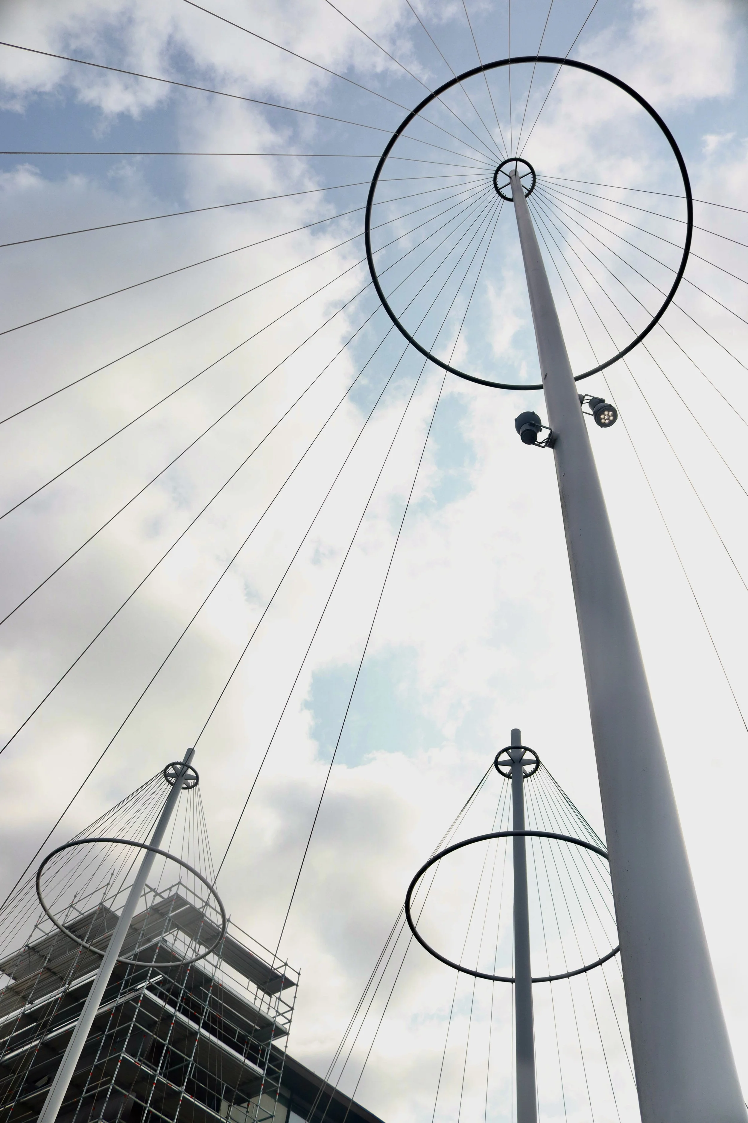 A low-angle view of tall white poles with circular metal rings at the top, linked by wires, against a partly cloudy sky; one pole has a couple of spotlights attached.