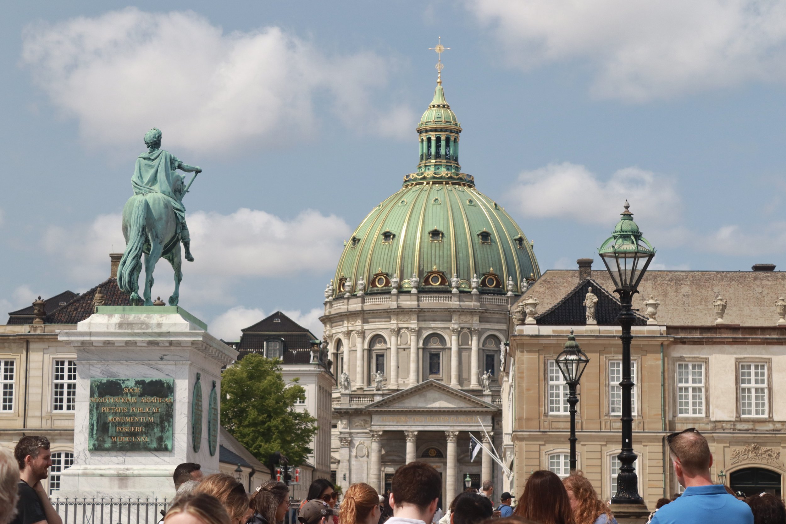 Crowds of people gathered in a city square with a large historic church featuring a green copper-domed roof in the background. A bronze equestrian statue is prominently displayed on a marble pedestal in the foreground.