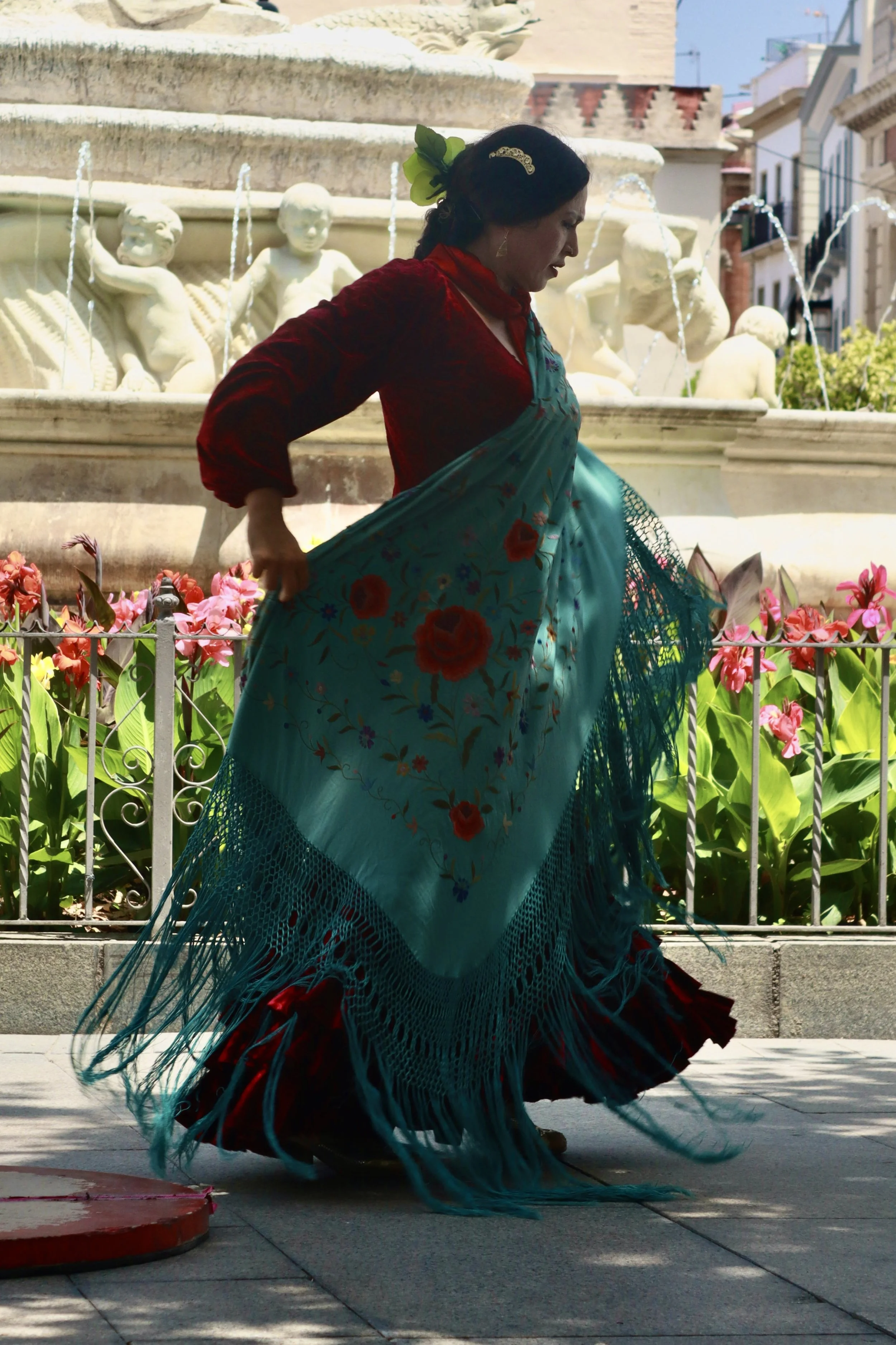 A woman in traditional Mexican attire dancing on a sidewalk in front of a fountain with sculptures and flowers.