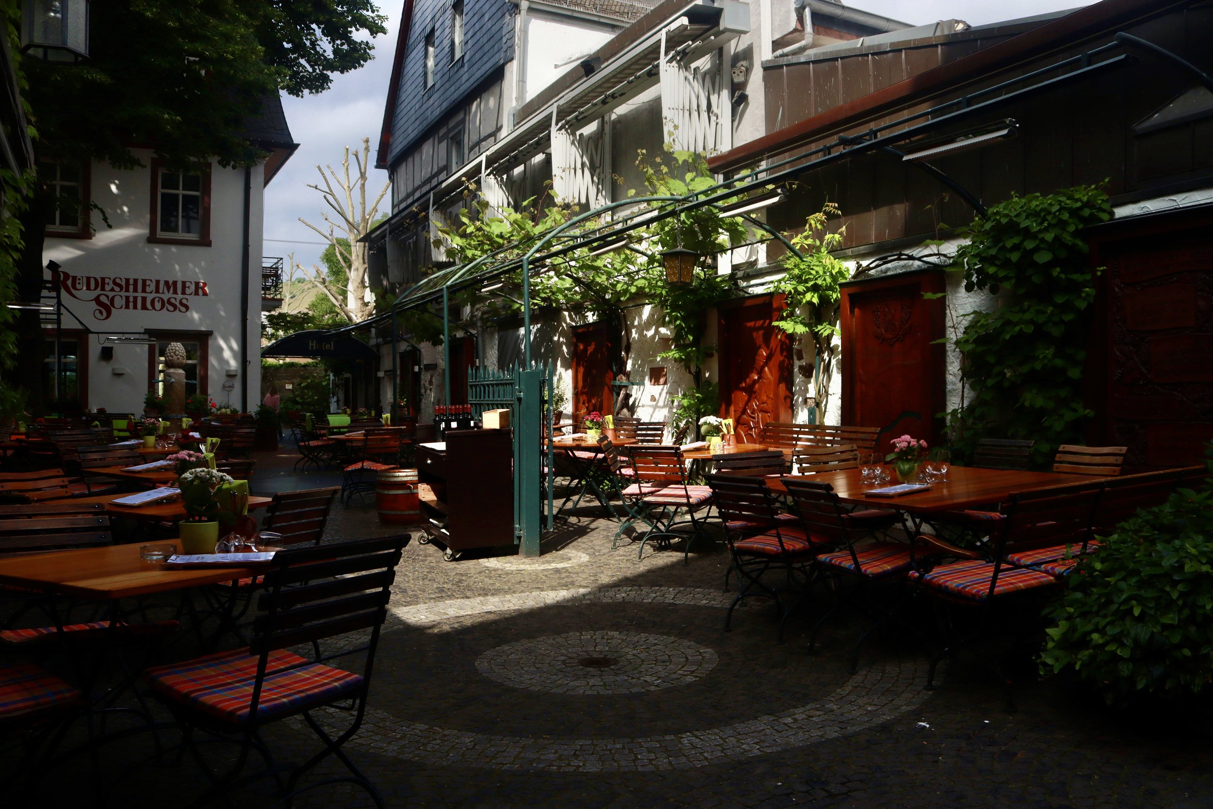 An outdoor dining area with wooden tables and striped cushioned chairs. The setting is shaded with green plants and climbing vines on the building walls. There are flowers and glasses on the tables, and it appears to be part of a restaurant or cafe i