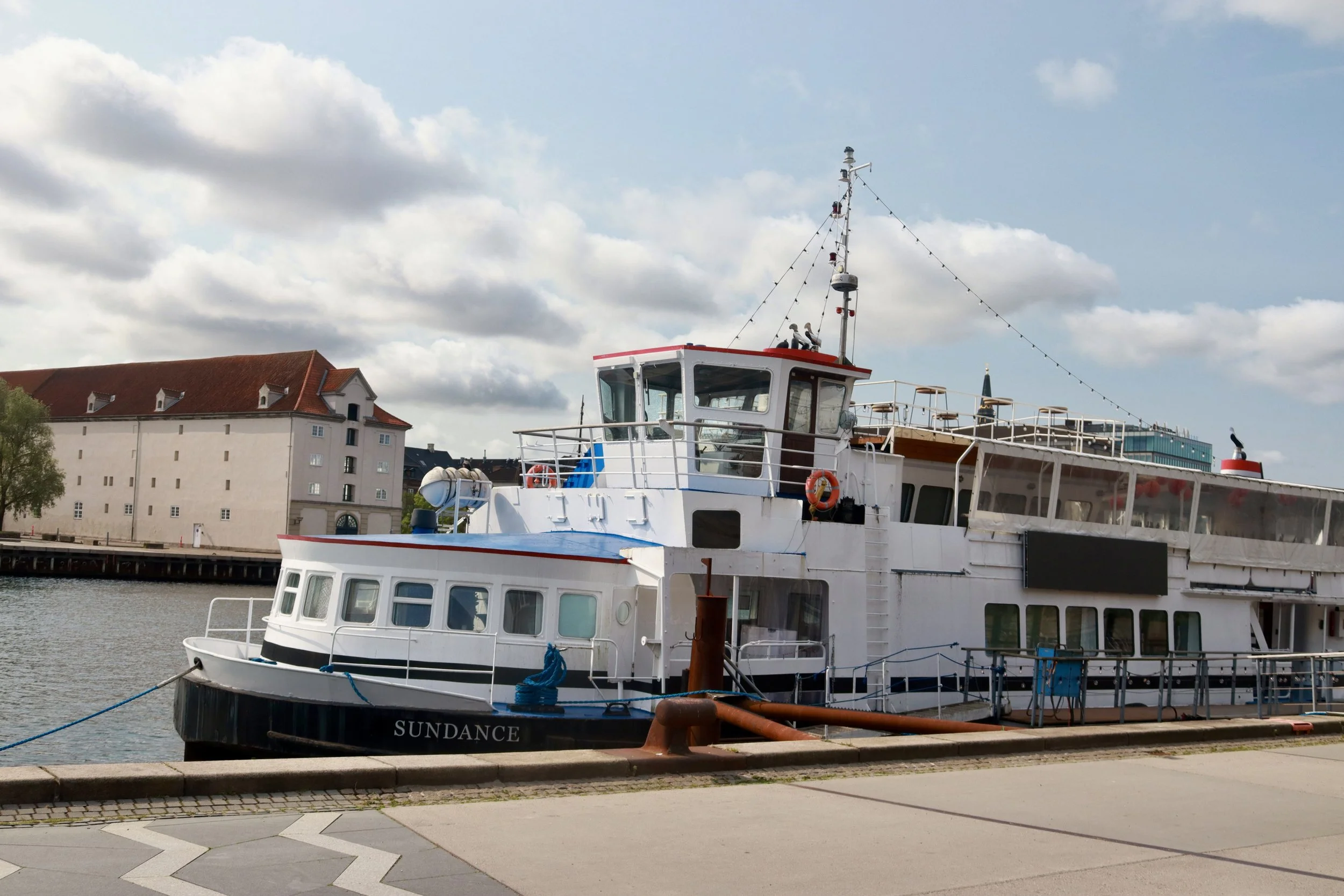 A white riverboat named Sundance docked at a pier, with buildings and a partly cloudy sky in the background.