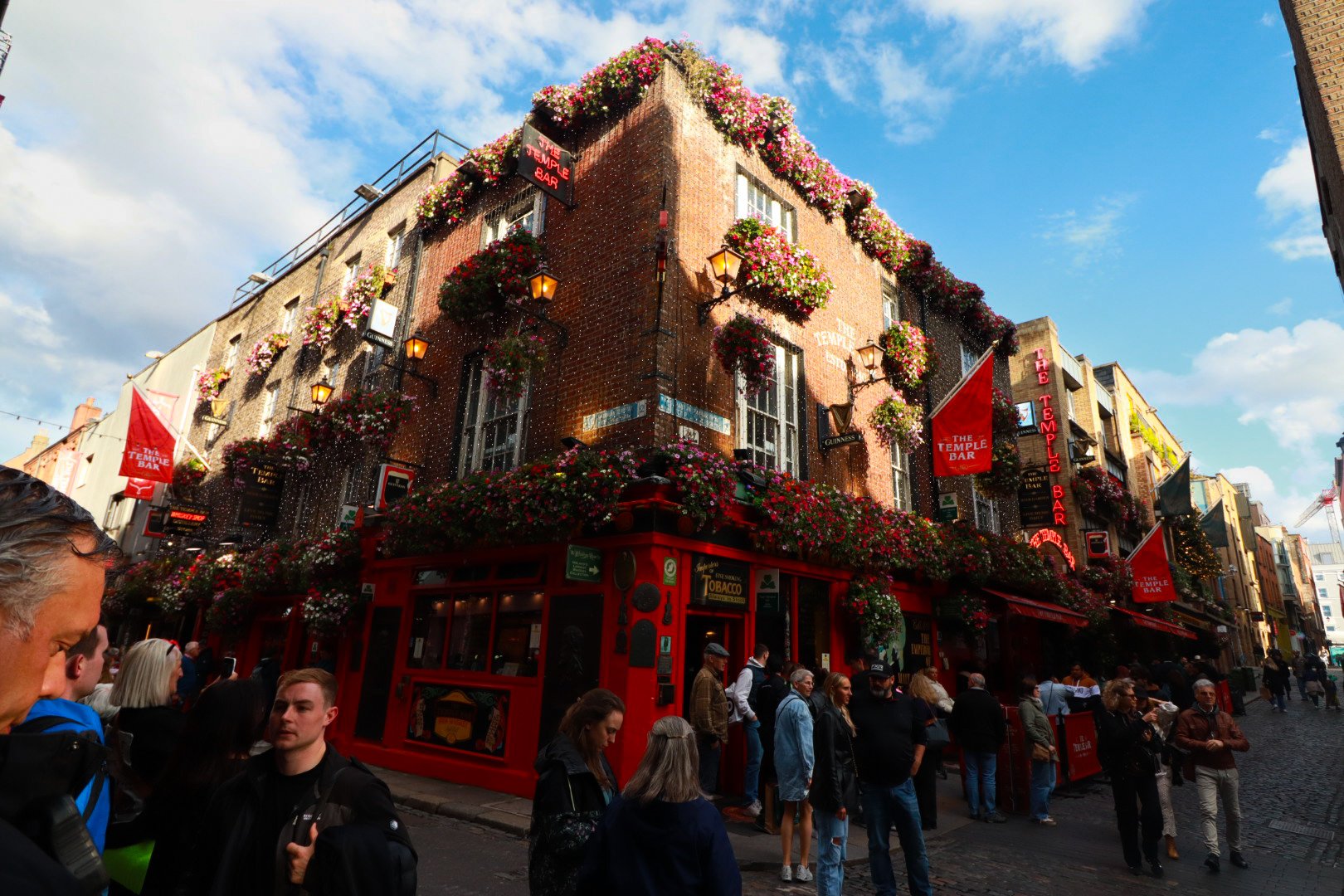 A corner pub named The Temple Bar in Dublin, Ireland, with red exterior, colorful flower arrangements, and neon signs, filled with people on a cobblestone street under a partly cloudy sky.