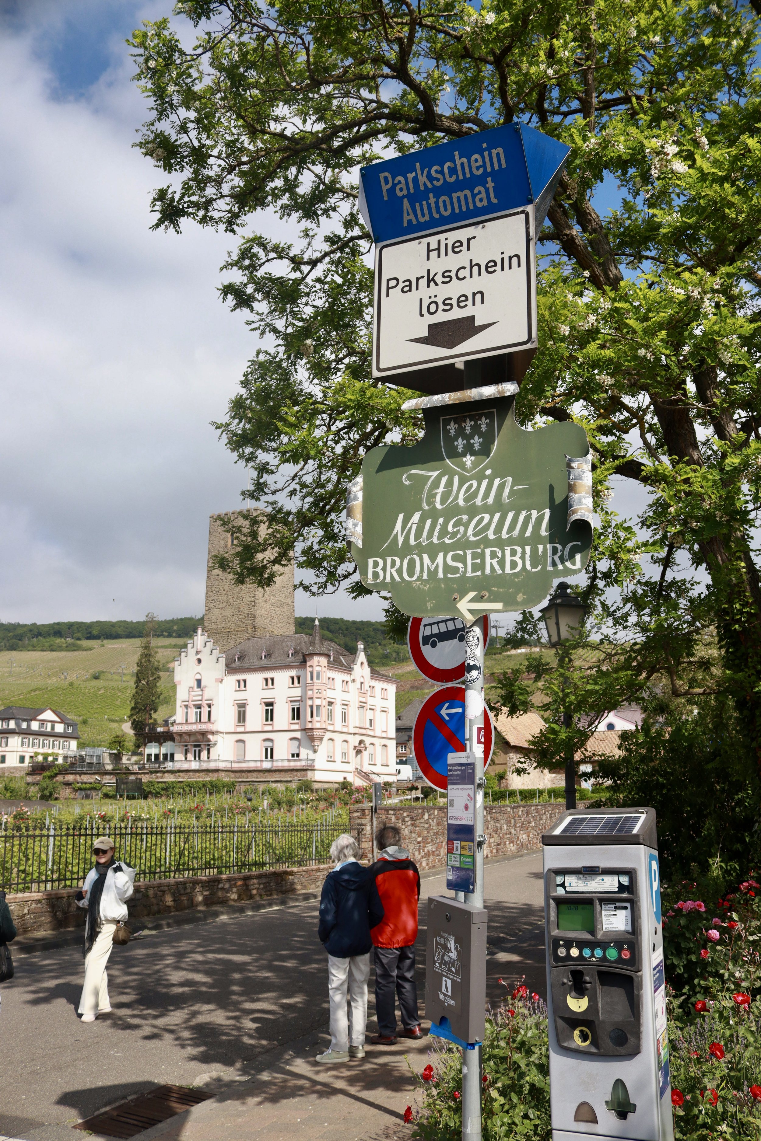 Street scene in a European town with a large signpost, a historic pink building, a castle tower in the background, and people walking. The signposts indicate parking automation, the Wein-Museum Bromserburg, and other parking rules.