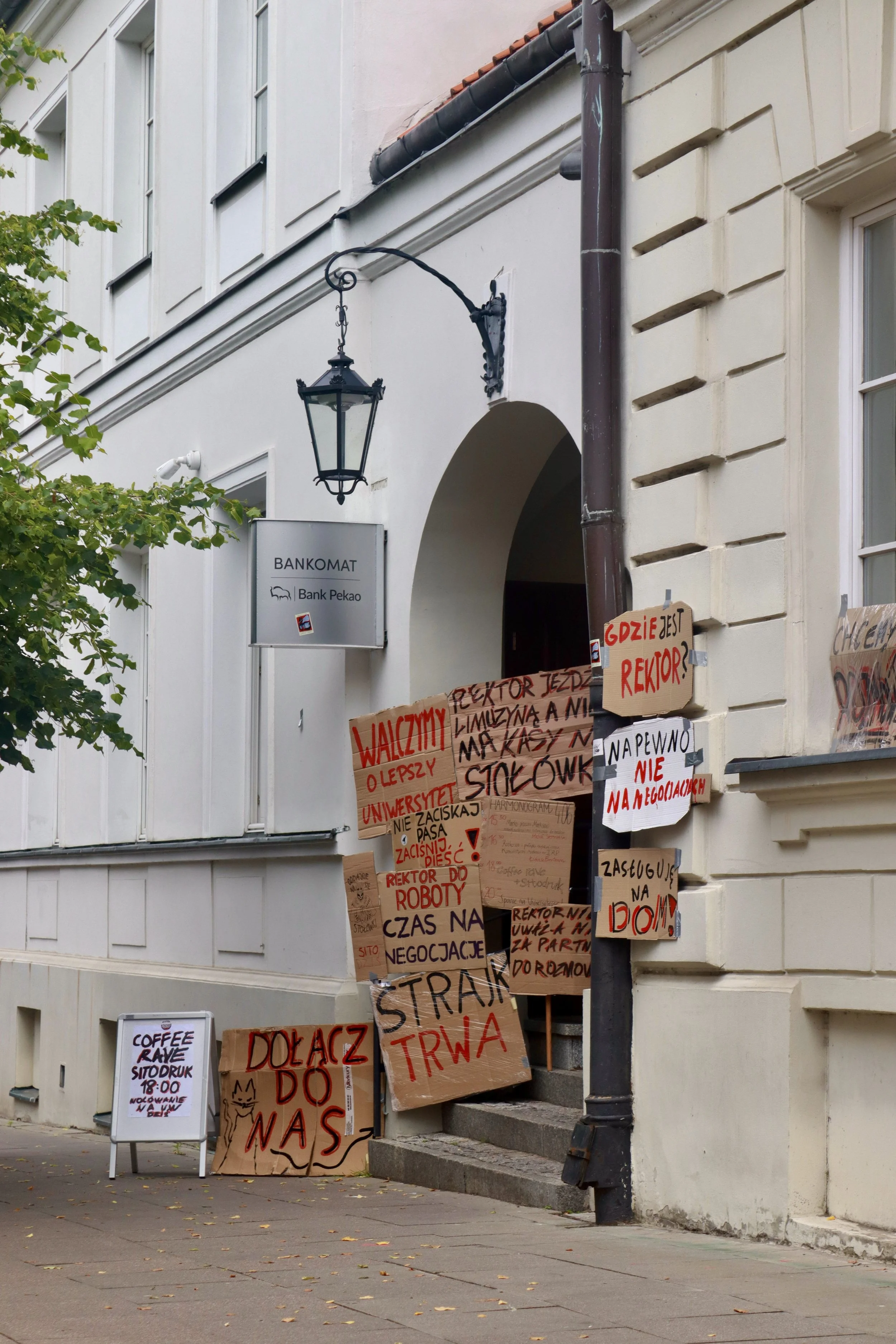 Protest signs on a city sidewalk with apartment building, bank sign, and street lamp in the background.