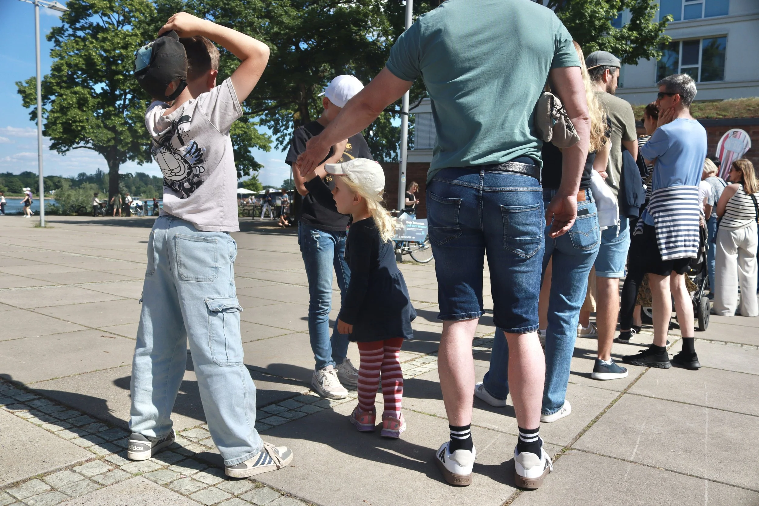 People queuing outdoors on a sunny day near a waterfront, with trees and a building in the background.