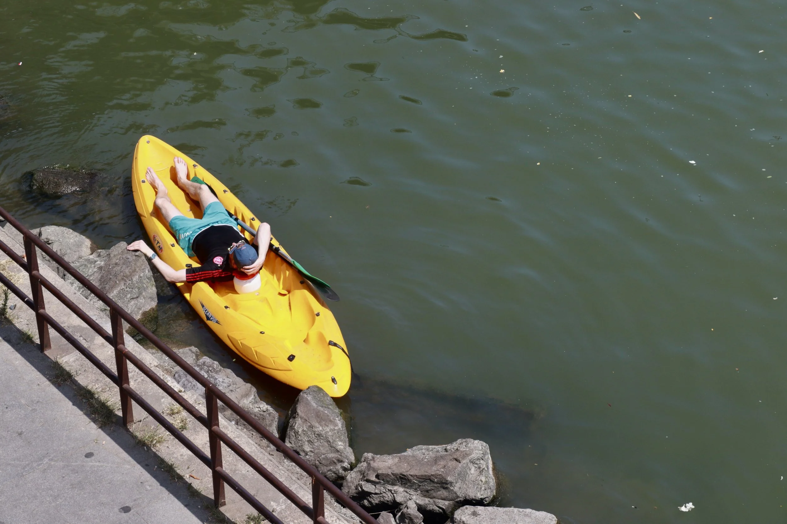Person lying on a yellow kayak resting on rocks beside a body of water.