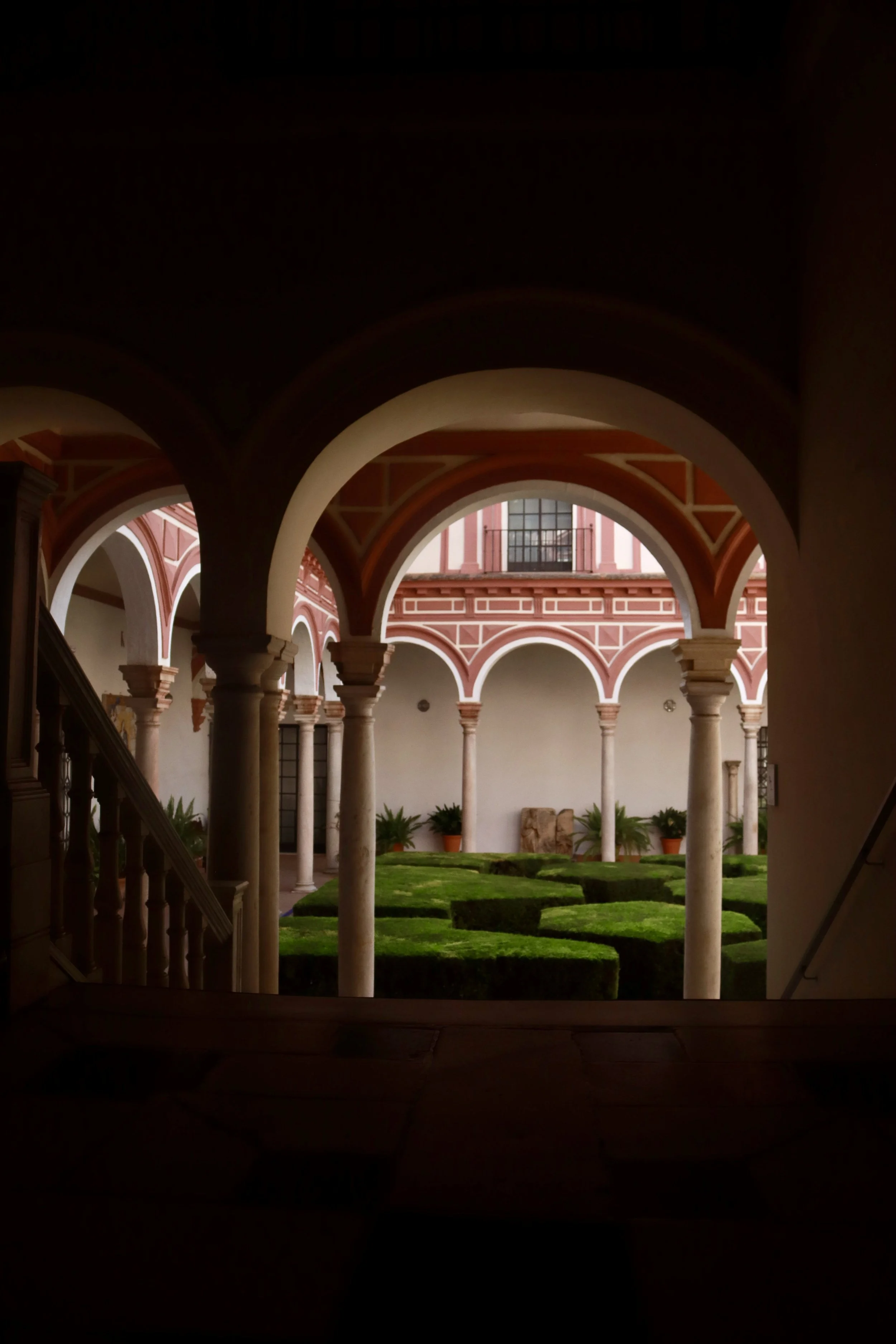 View of a courtyard through arches, with trimmed green bushes, potted plants, and a pink building with white trim in the background.