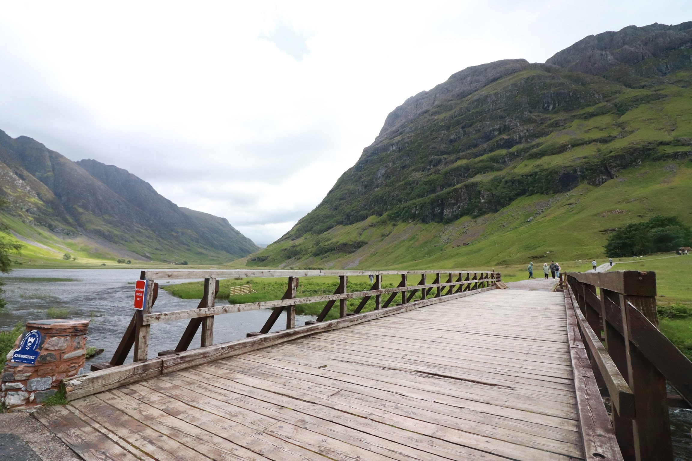 A wooden bridge over a river with green mountains in the background and a cloudy sky.