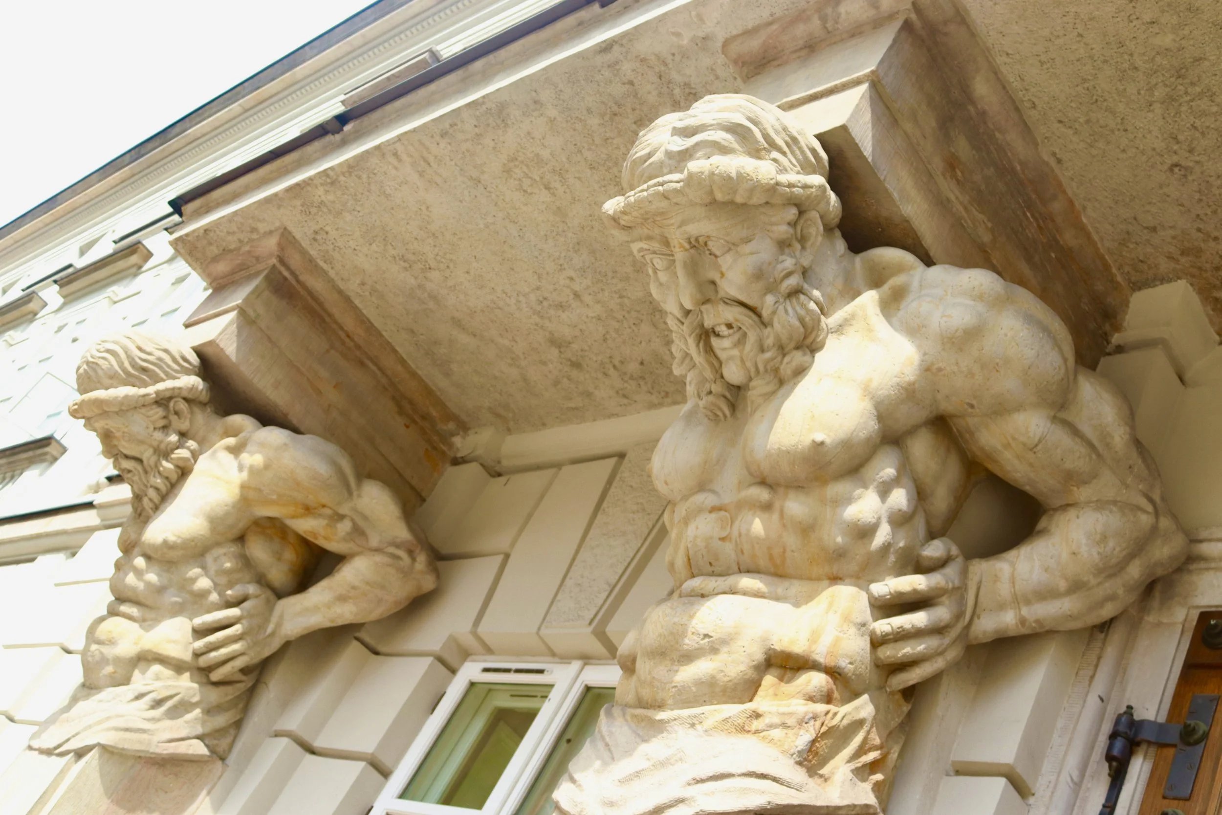 Close-up of two stone sculpted male busts with muscular torsos attached to the exterior of a building, showing detailed facial features and beards.