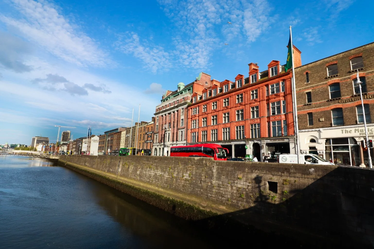 Cityscape along a river with historic brick buildings, a red bus, and a blue sky with scattered clouds.