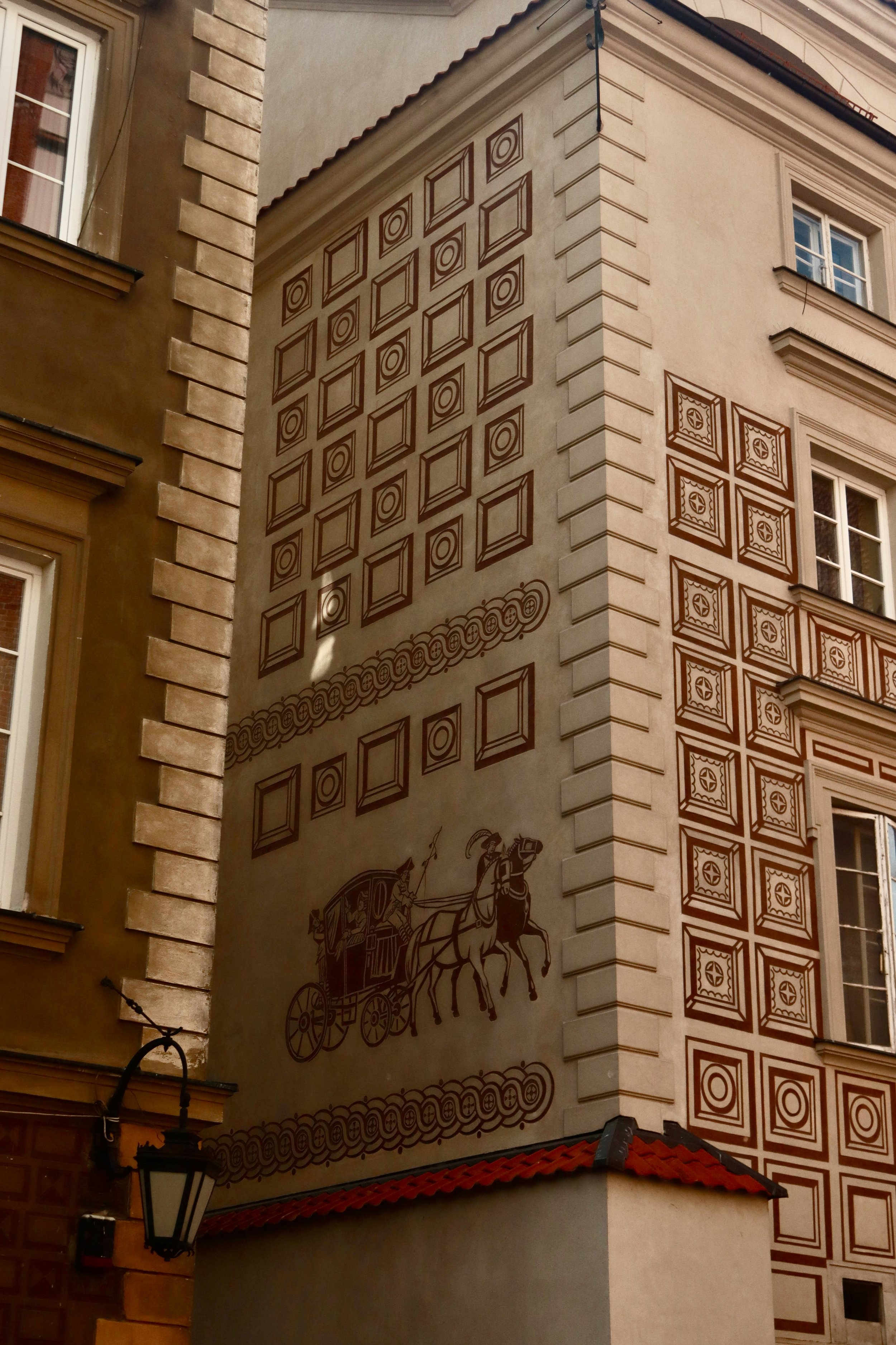 A decorative mural on the side of a building displaying historical imagery of a horse-drawn carriage and people, with patterned square designs and circular motifs.