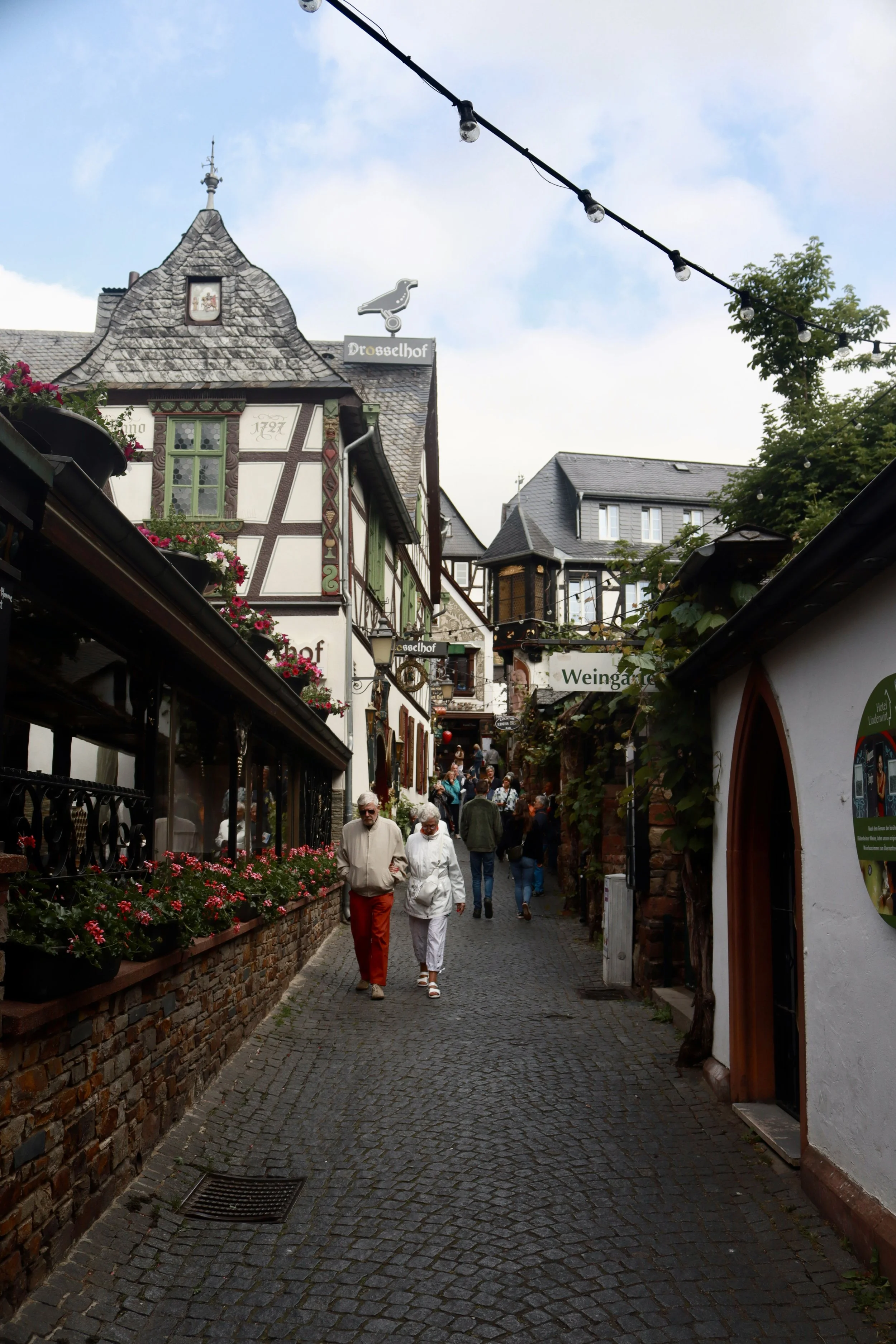 A cobblestone street in a quaint German town with half-timbered buildings, flower boxes, and pedestrians walking under a partly cloudy sky.