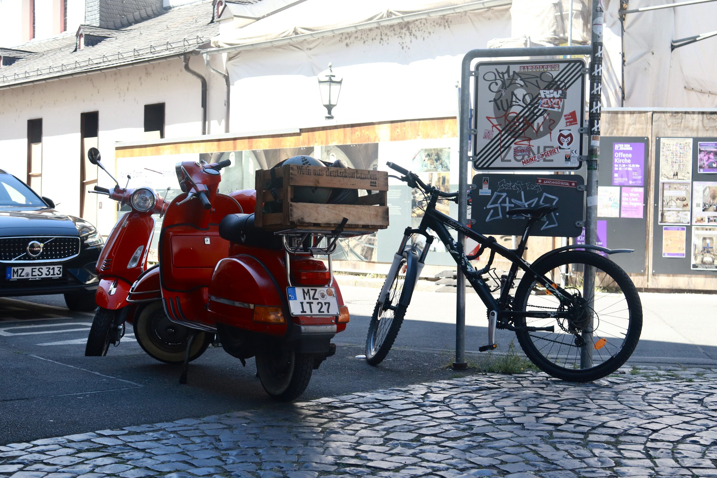 A red vintage scooter with a wooden crate on the back, parked next to a black mountain bike locked to a post on a cobblestone street.