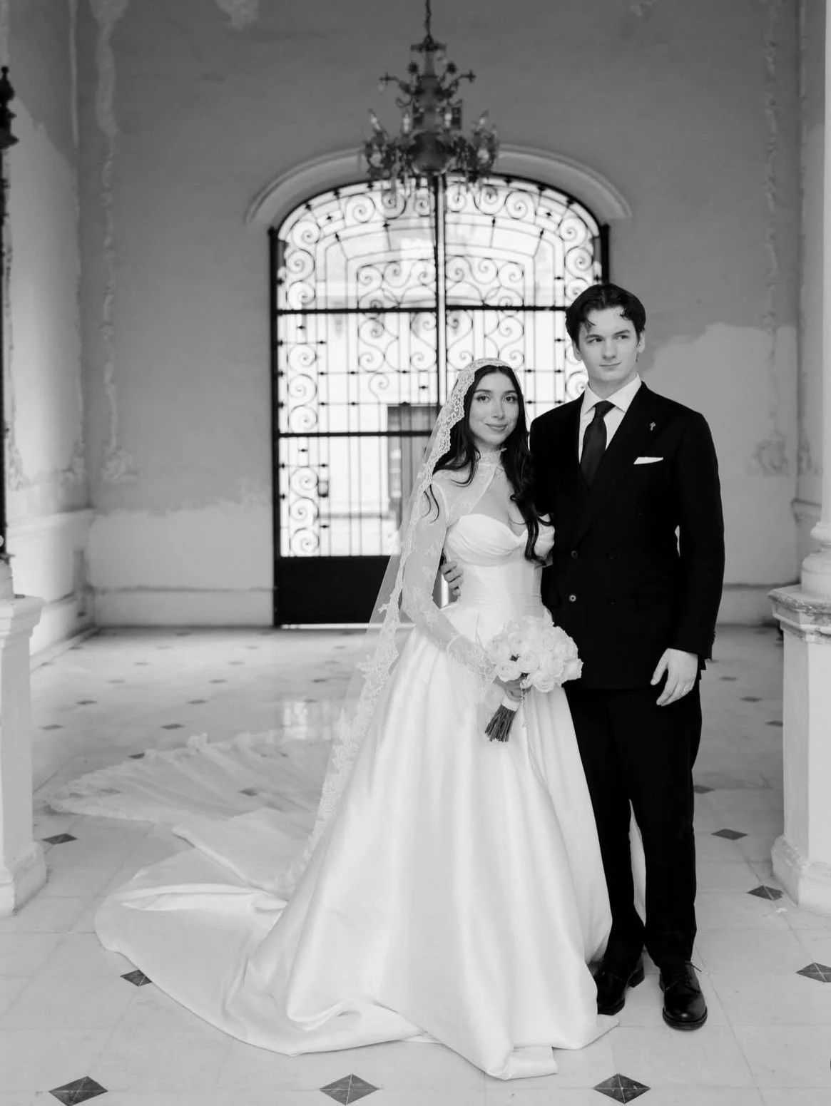 Black and white photo of a bride and groom standing inside a historic building with ornate walls and a decorative wrought iron gate.